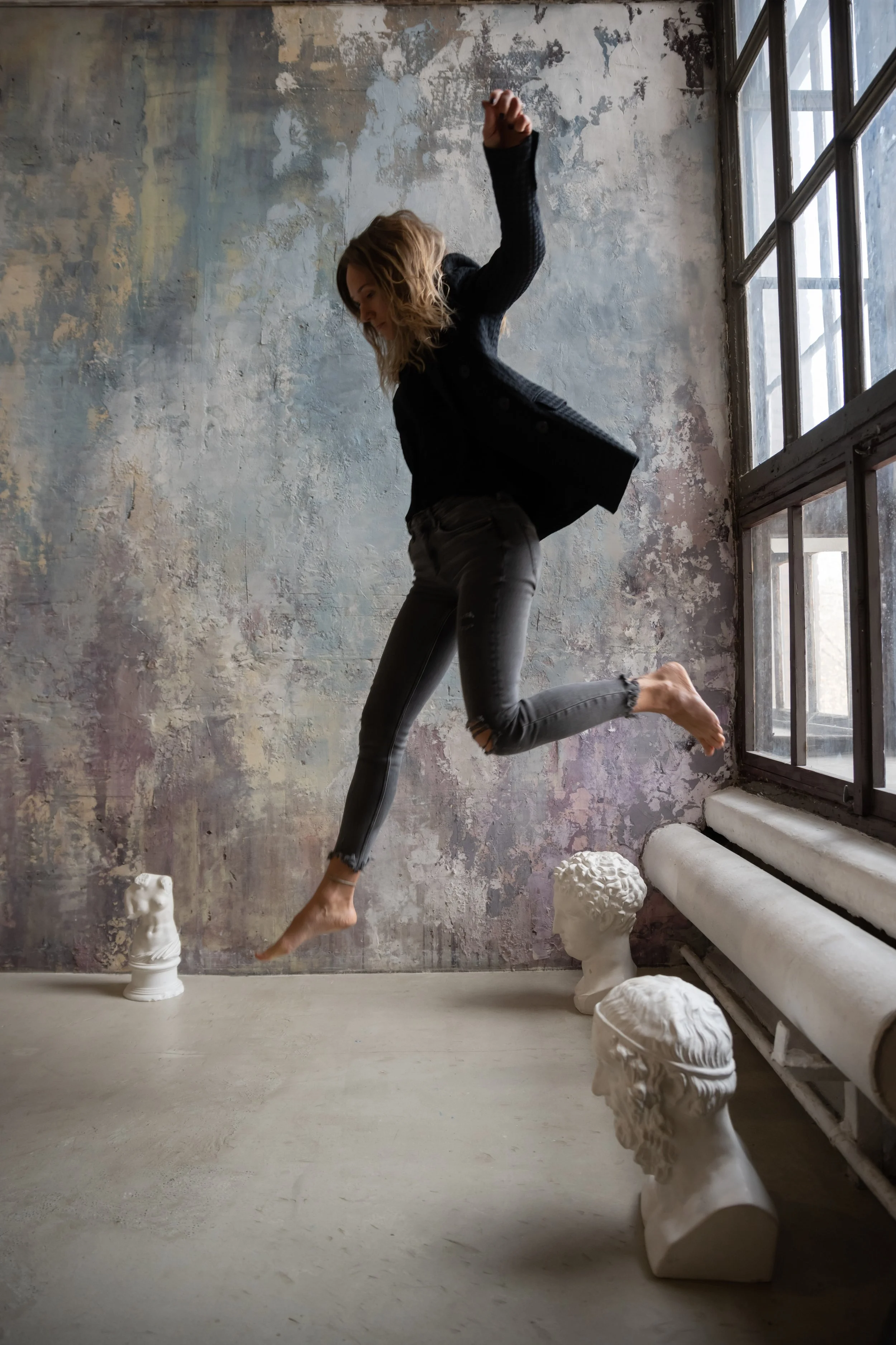 Une femme en saut dans une pièce avec un mur en béton et des statues de bustes romains en plâtre.