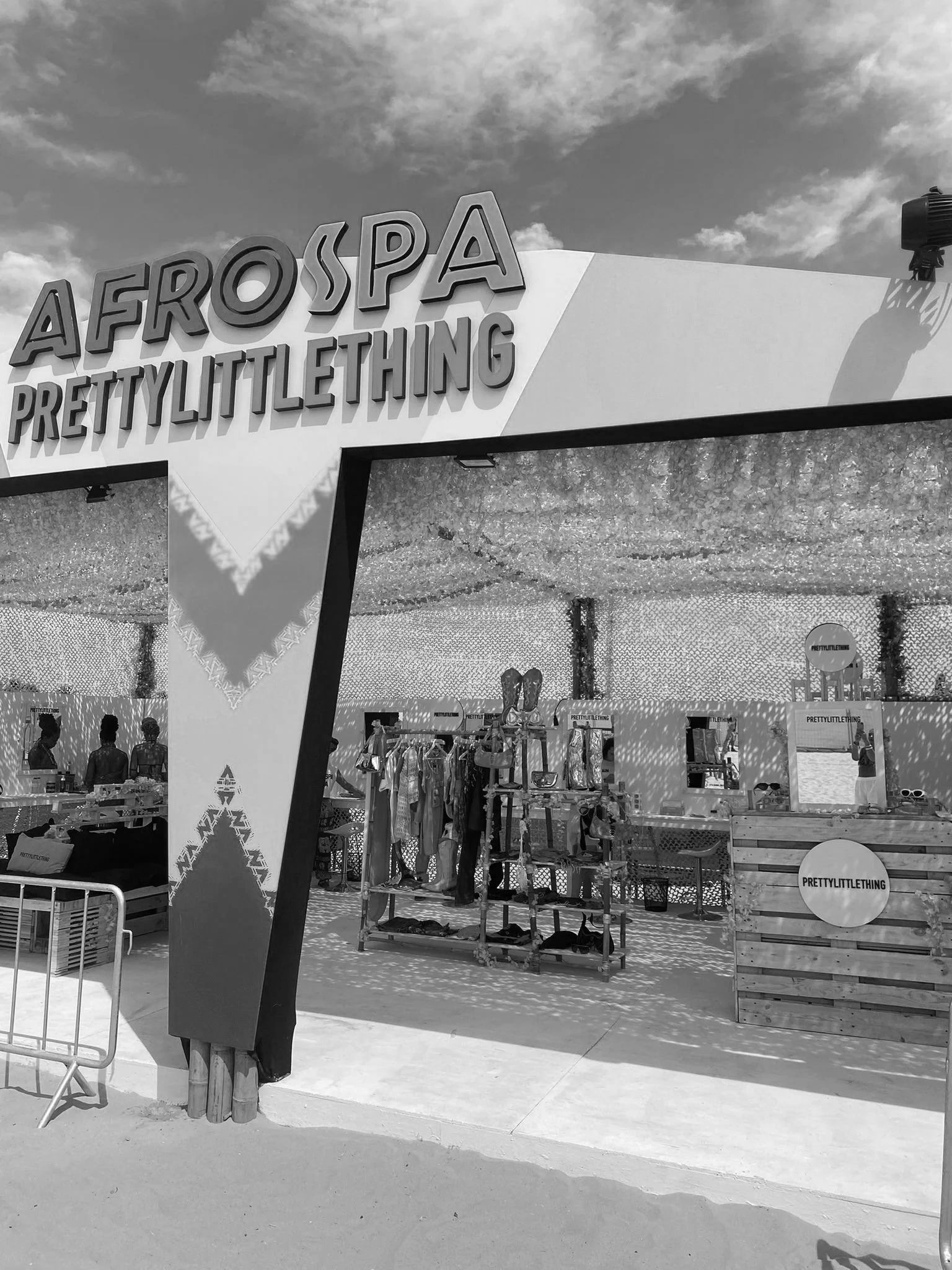 An outdoor store with a large sign that reads 'A F R O S P A PRETTYLITTLETHING'. The store sells clothing and accessories, with racks and display tables, and a canopy covered with lights.