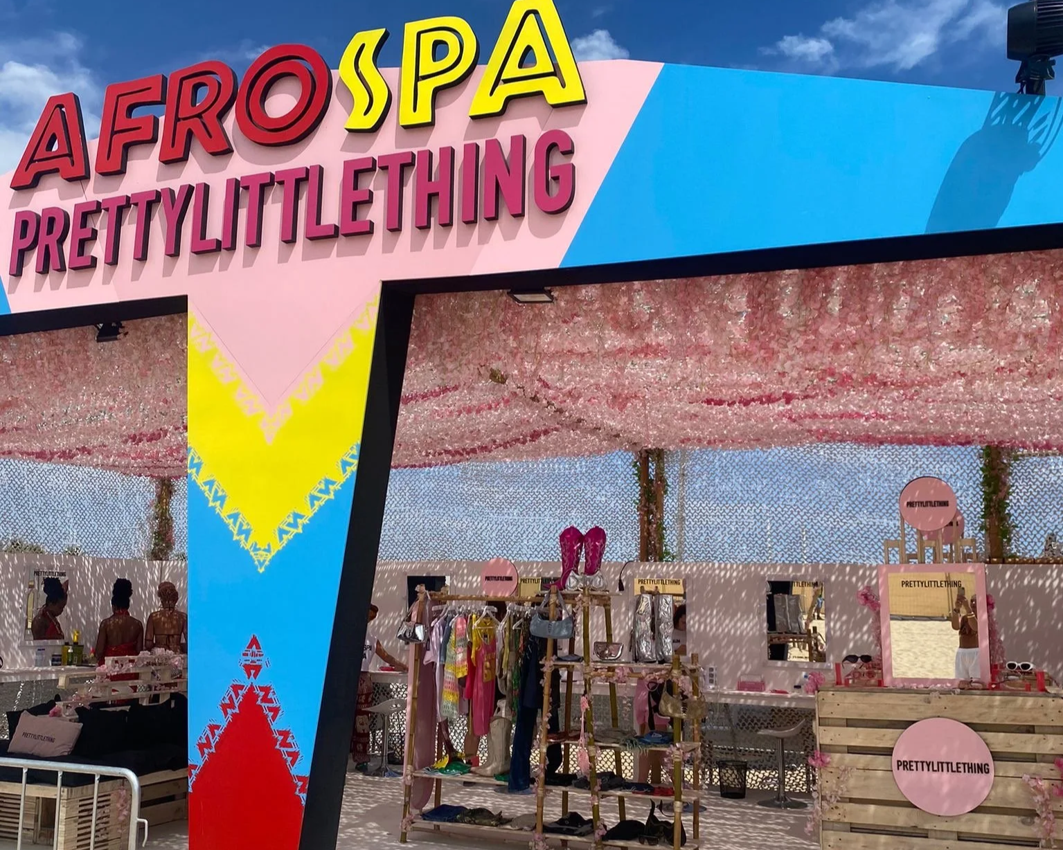A colorful outdoor stall at an event with sign reading 'AFRO SPA PRETTY LITTLE THING'. The stall has pink, blue, yellow, and red decorations, with clothing and accessories displayed on racks and tables. Pink floral decorations hang above the booth.