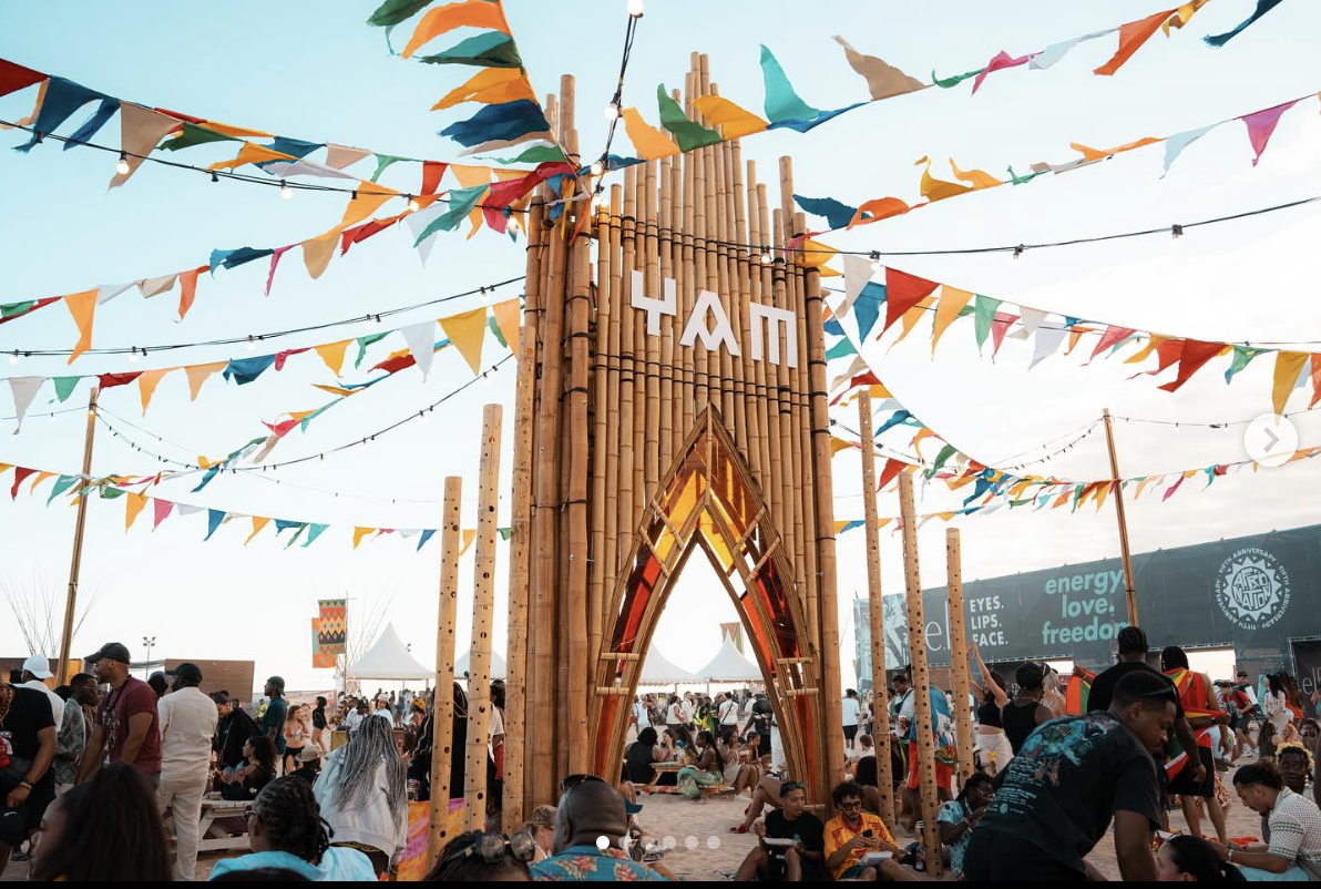 A festival scene with a large bamboo entrance structure decorated with colorful triangular flags strung across the sky, with many people gathered around, some sitting and others standing, in front of tents and banners at an outdoor event.