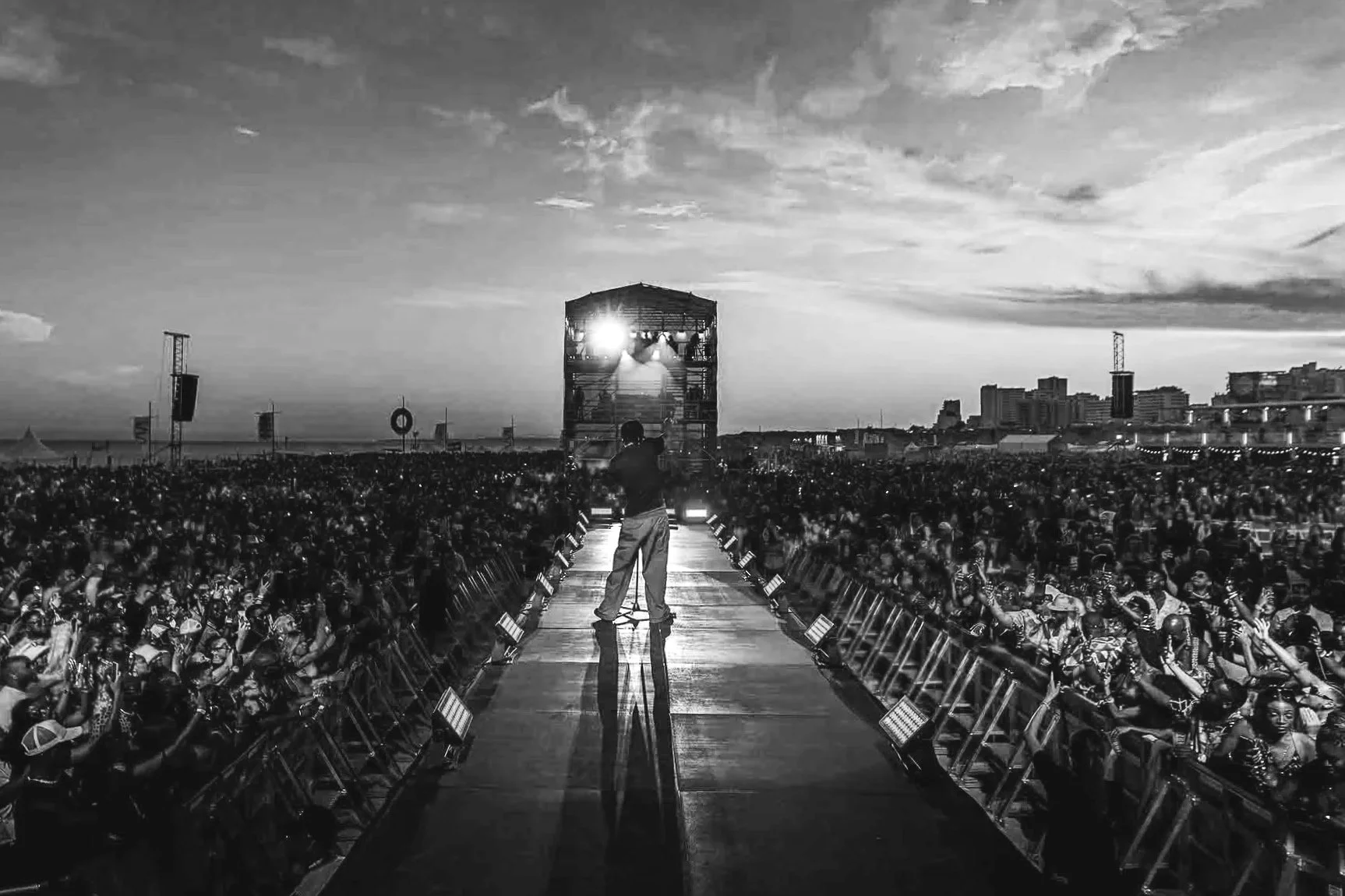 Black and white photo of a performer on stage at sunset with a large audience, city skyline in the background.