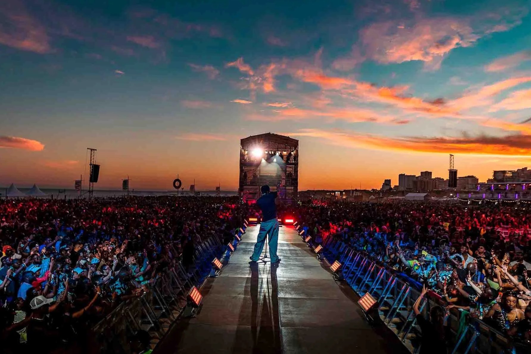 A performer on stage at an outdoor concert during sunset, with a large crowd of attendees in front, and a city skyline in the distance.