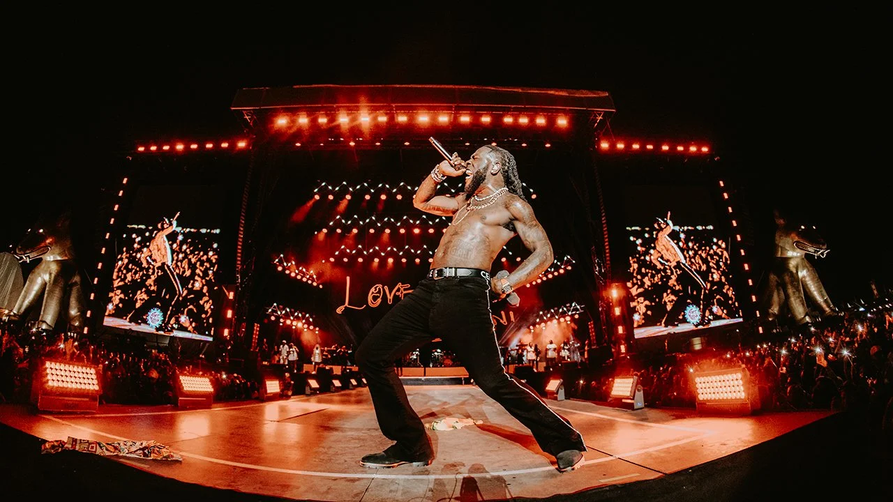Performer singing shirtless on stage with red lighting and large screen displays, surrounded by audience at night.