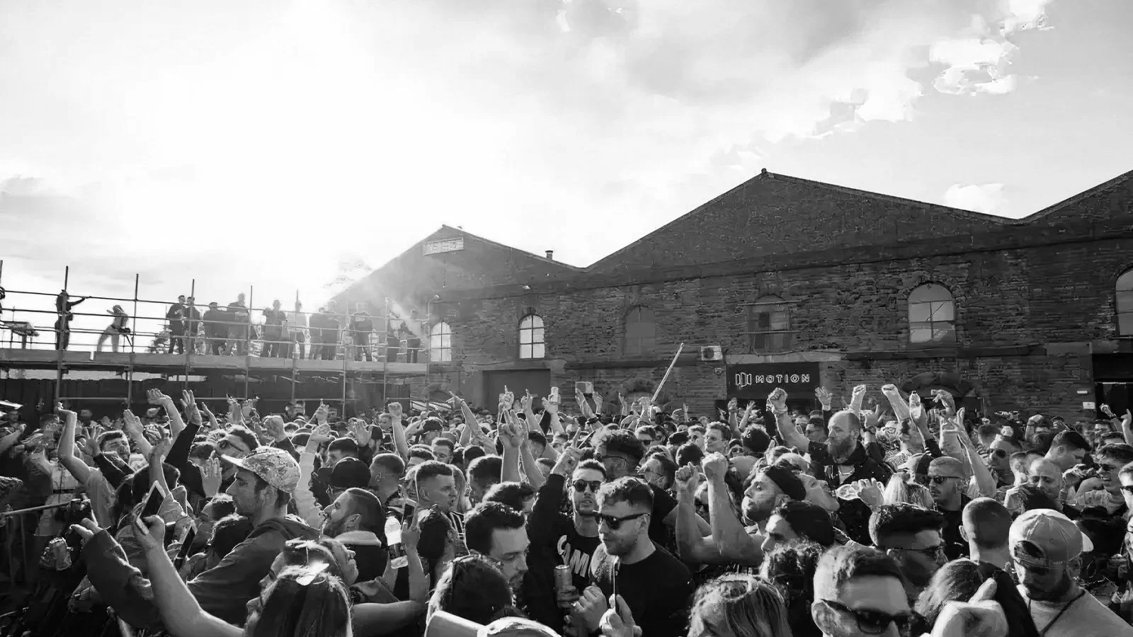 Black-and-white photograph of a large crowd of people attending an outdoor event, with some protesters on scaffolding and a brick building in the background.