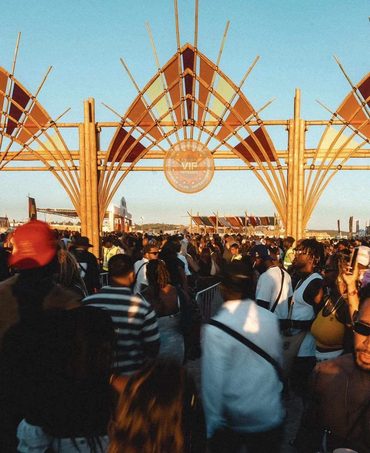 Crowd of people waiting in line at an outdoor event under a large wooden entrance arch with a VIP sign.