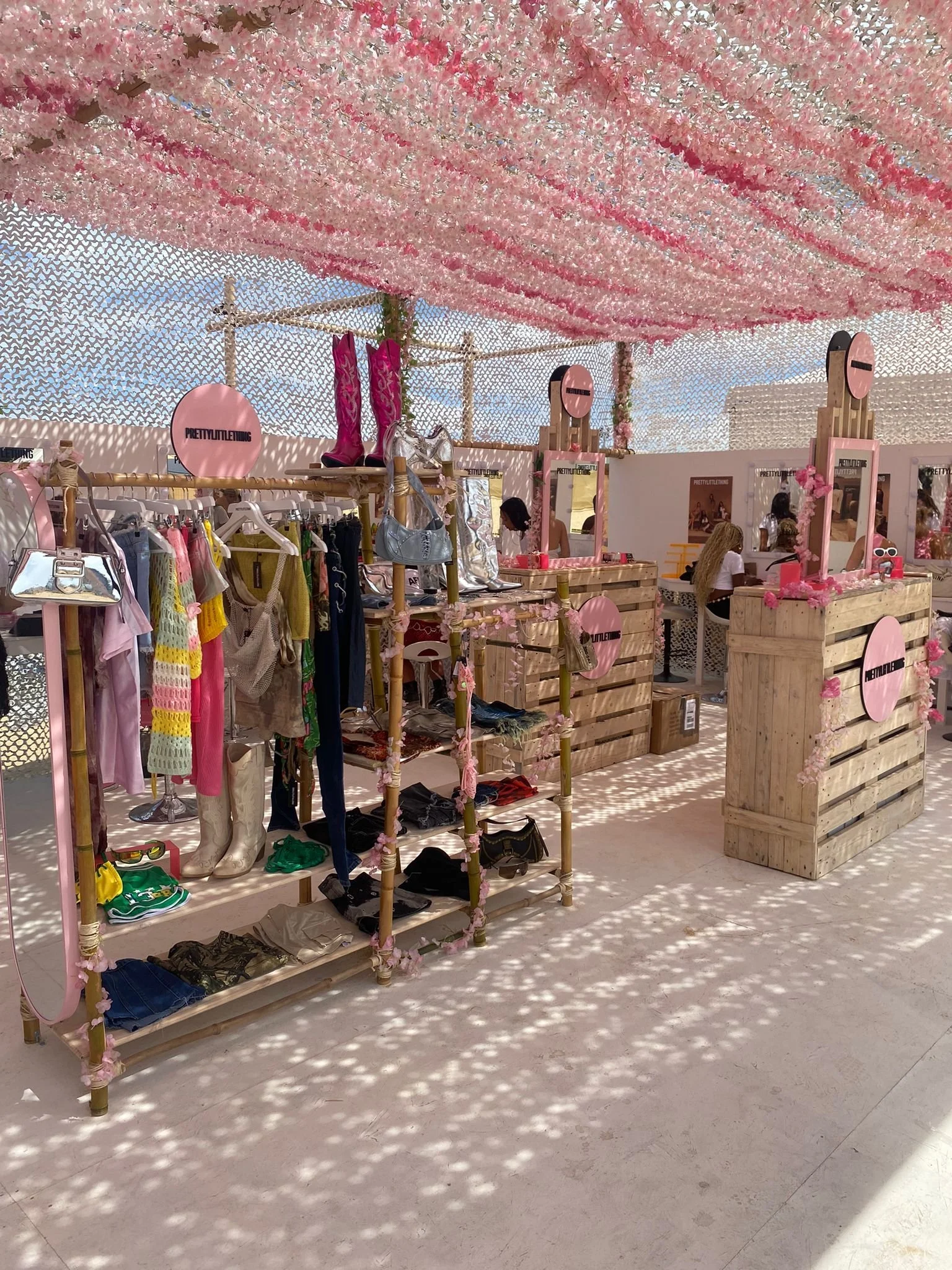 A clothing and accessories booth at an outdoor market decorated with pink and white flowers on the ceiling and pink accents, displaying various items like shoes, bags, and clothes.