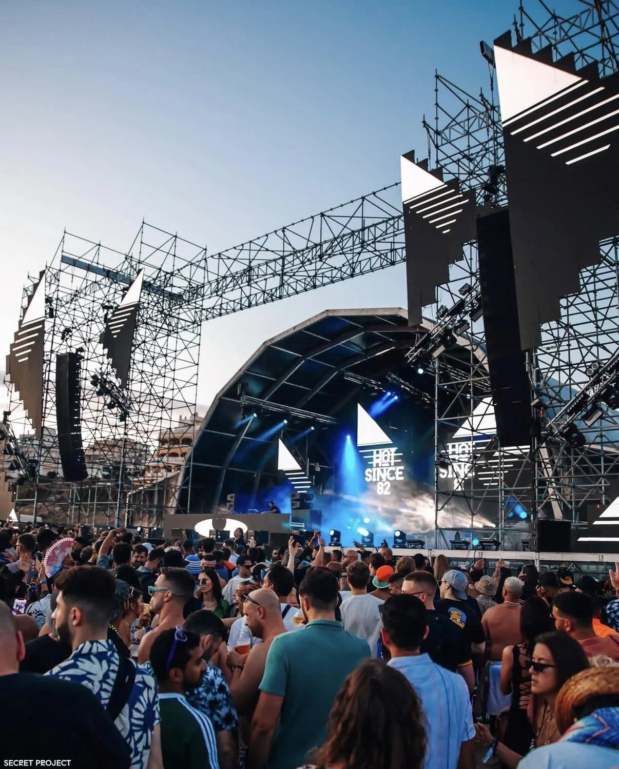 Crowd at an outdoor music festival stage during sunset, with lights and screens on the stage.