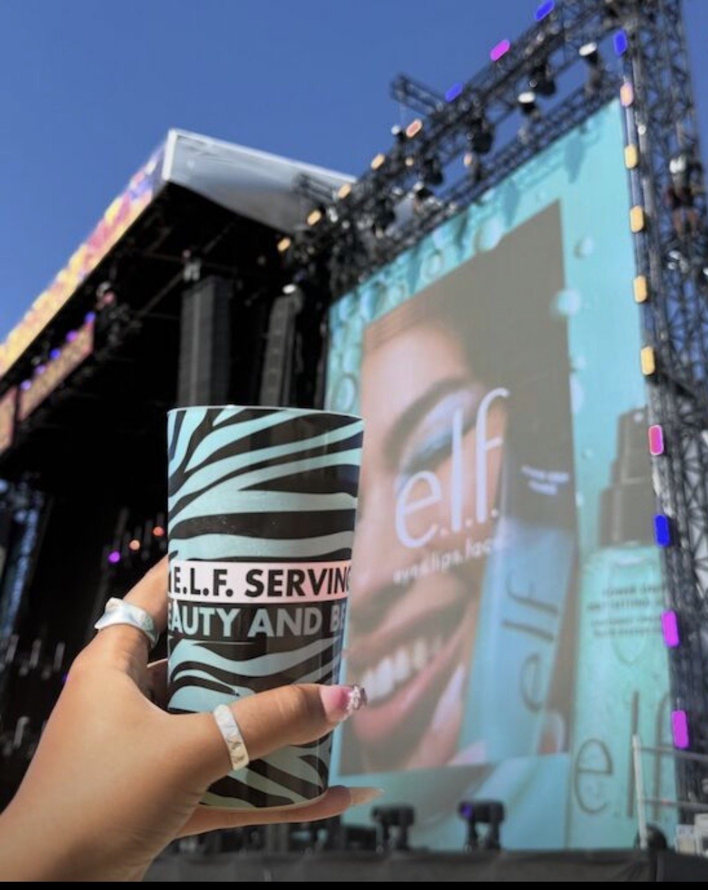A person holding a black and white zebra stripe patterned tumbler with the words 'SELF. SERVING. BEAUTY AND BE' in front of a large outdoor stage with a screen showing a close-up of a face and an e.l.f. cosmetics product.