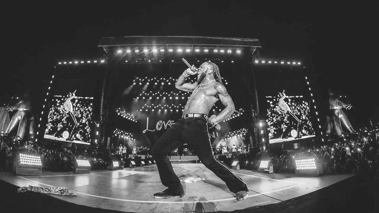 A shirtless male performer singing into a microphone on stage with bright lights and large screens showing his image in the background.