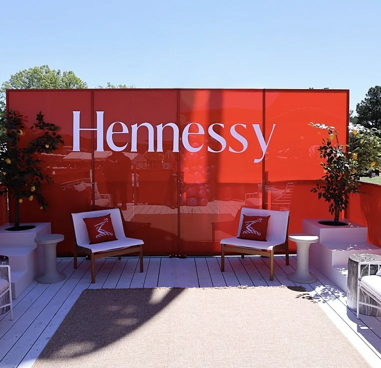 Outdoor lounge with red Hennessy signage, white chairs with pillows, potted plants, and side tables on a wooden deck.