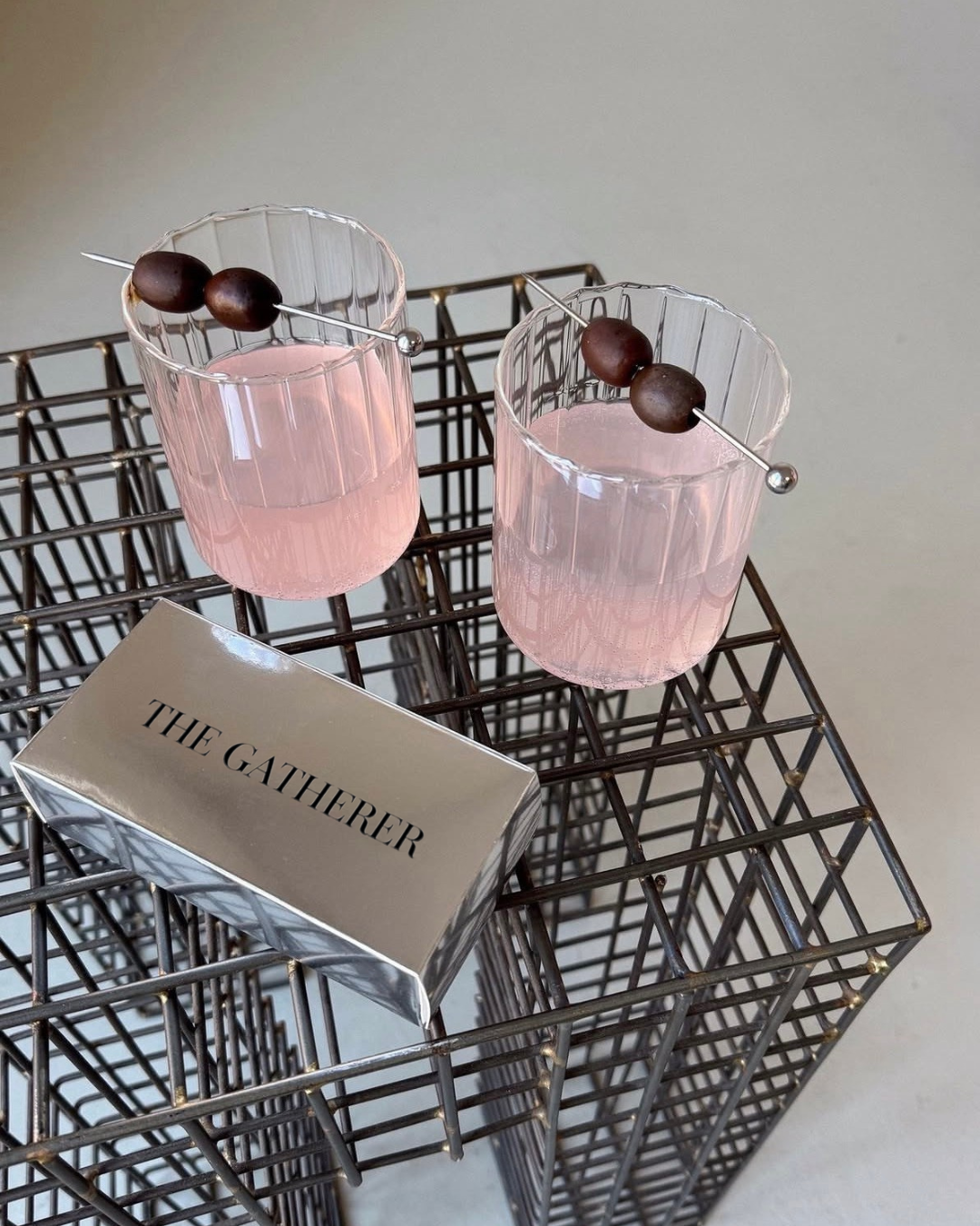 Two glasses with pink drinks topped with skewer of dark round olives, placed on a metallic wire basket next to a silver box labeled 'The Gatherer'.