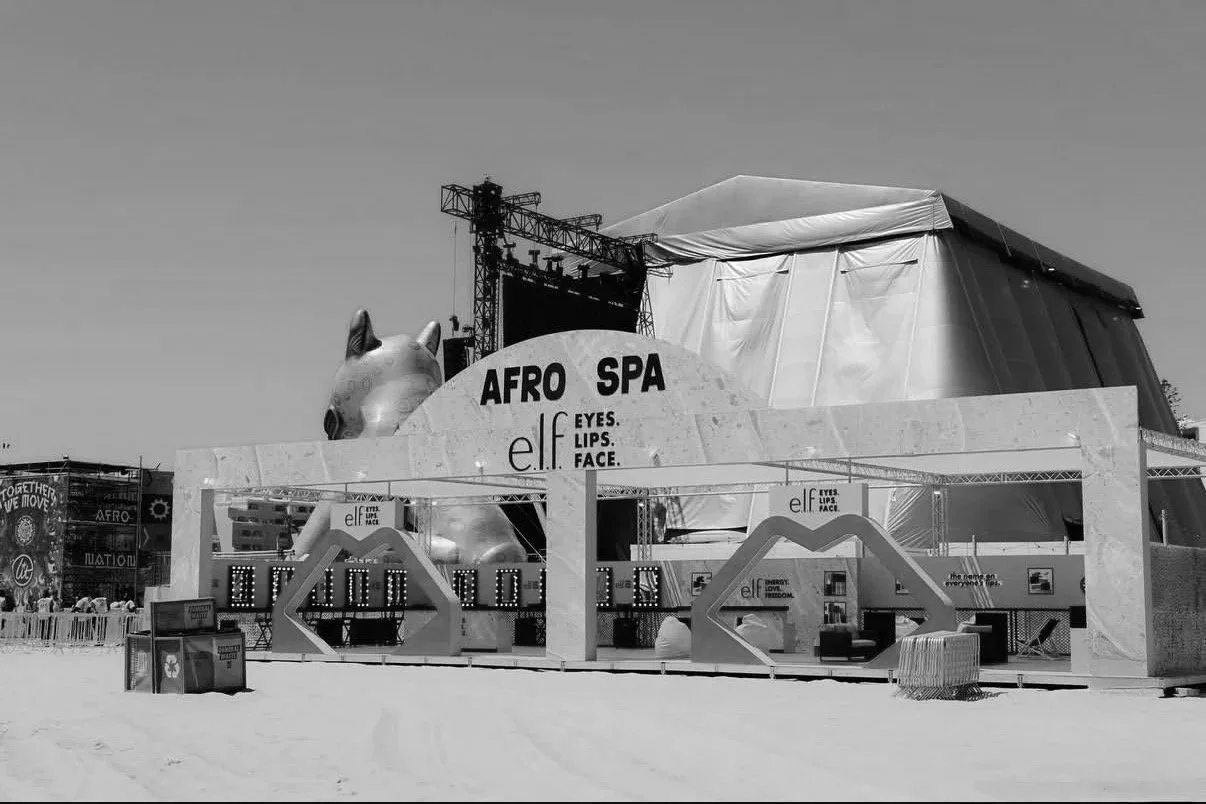 Empty festival booth for the Afro Spa promoting elf cosmetics, with a large horse head sculpture and stage setup in the background at an outdoor event.