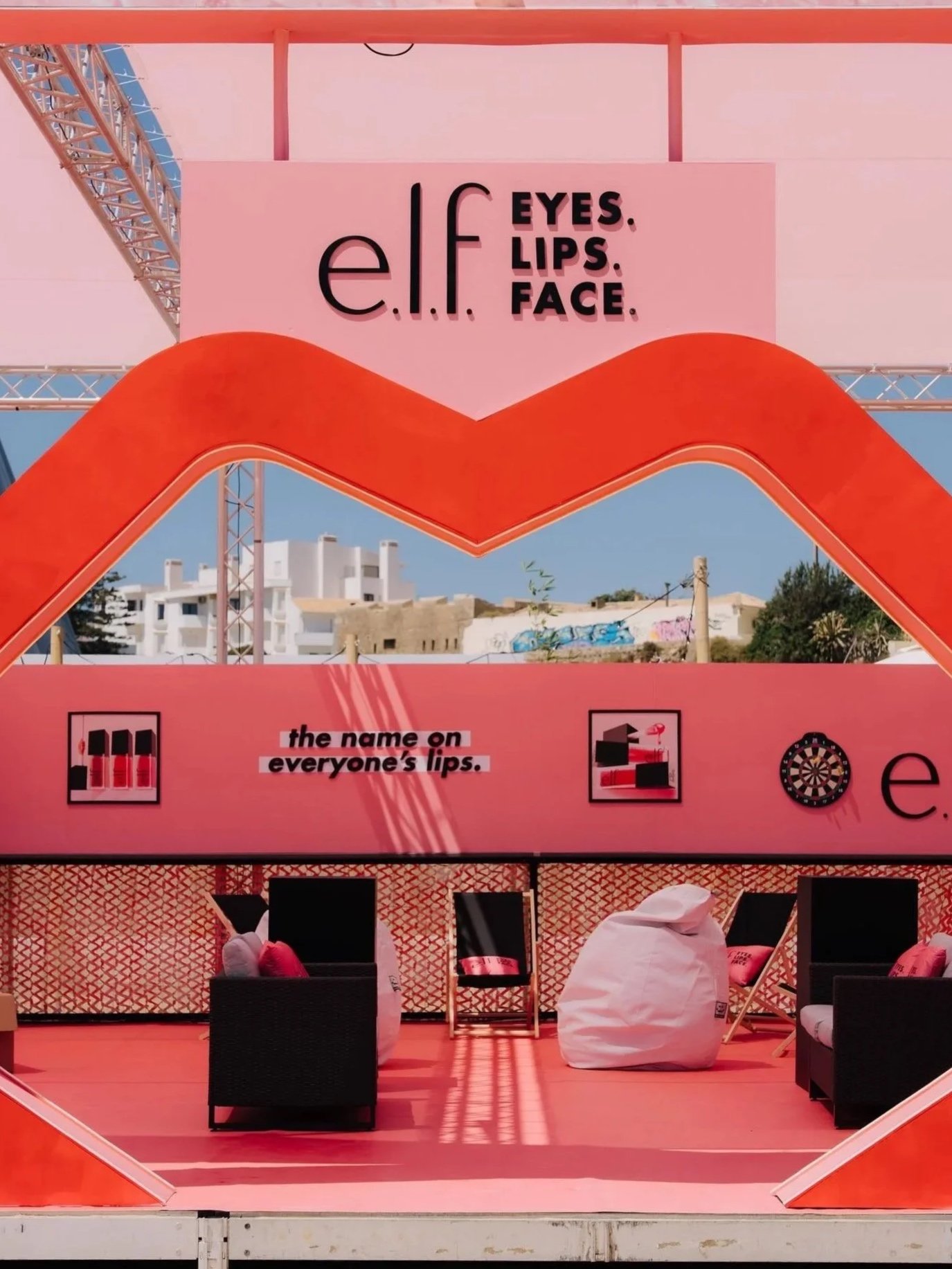 Display booth with pink and orange decorations promoting elf cosmetics, featuring signs about eyes, lips, and face, with seating area including black chairs and bean bags.