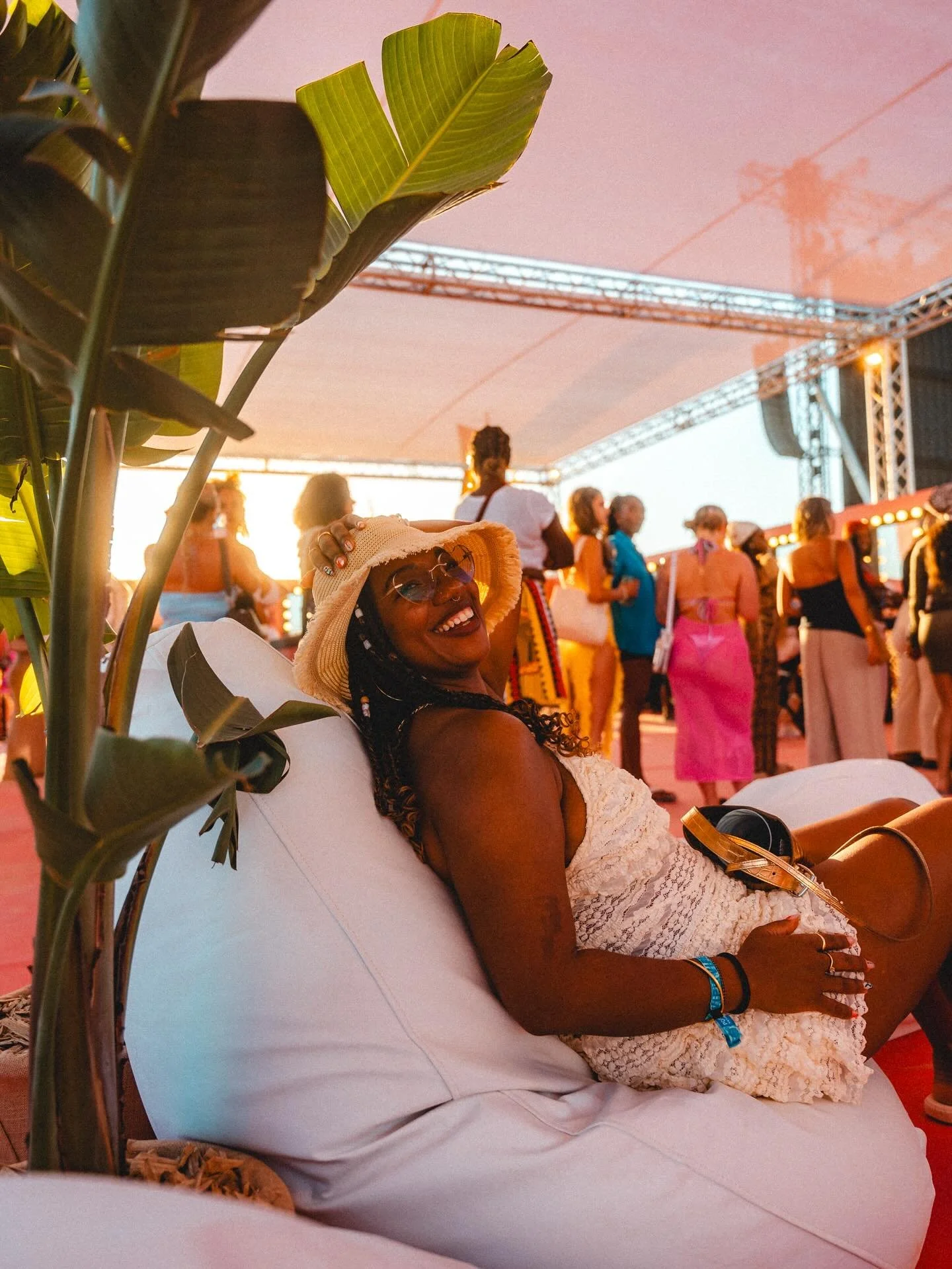 A woman wearing a sunhat and glasses, smiling and relaxing on a lounge chair at an outdoor event with a crowd of people in the background during sunset.