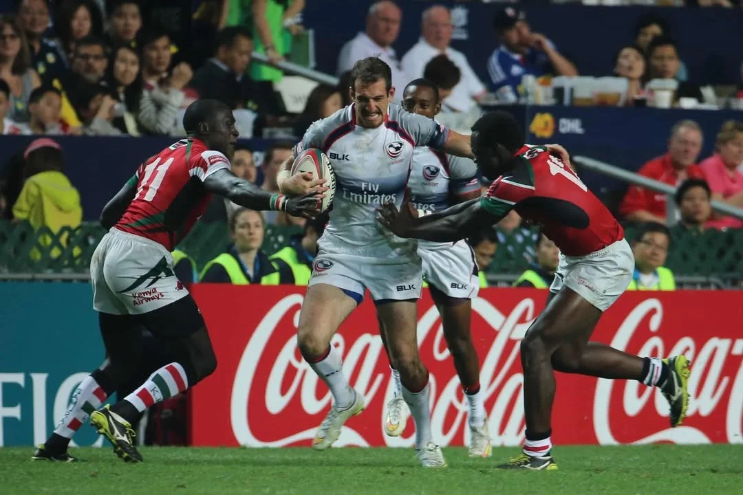 Rugby players in action during a game; one player in white is holding the ball, surrounded by players in red attempting to block or tackle.