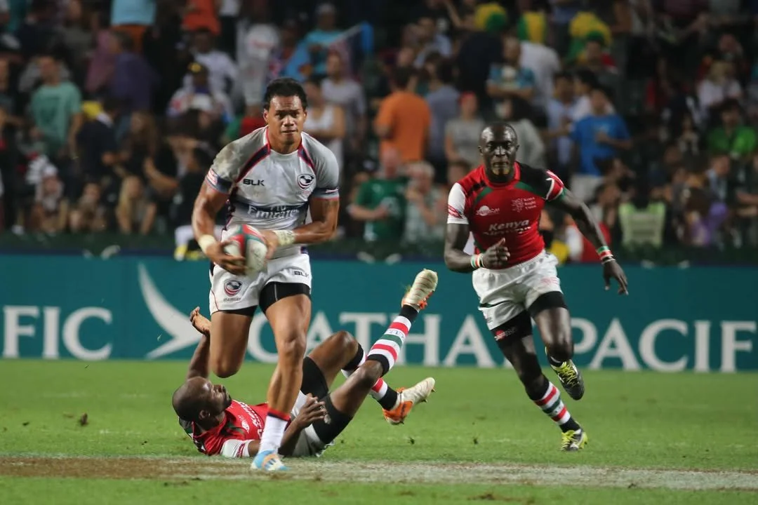 Rugby players in action during a game, with one player holding the ball and two players on the ground attempting to tackle him on a grass field, spectators in the background.