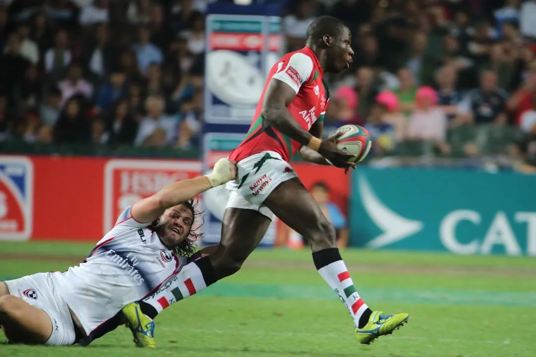 A rugby player in a red jersey runs with the ball while a player in a white jersey with red and black accents attempts to tackle him from behind.