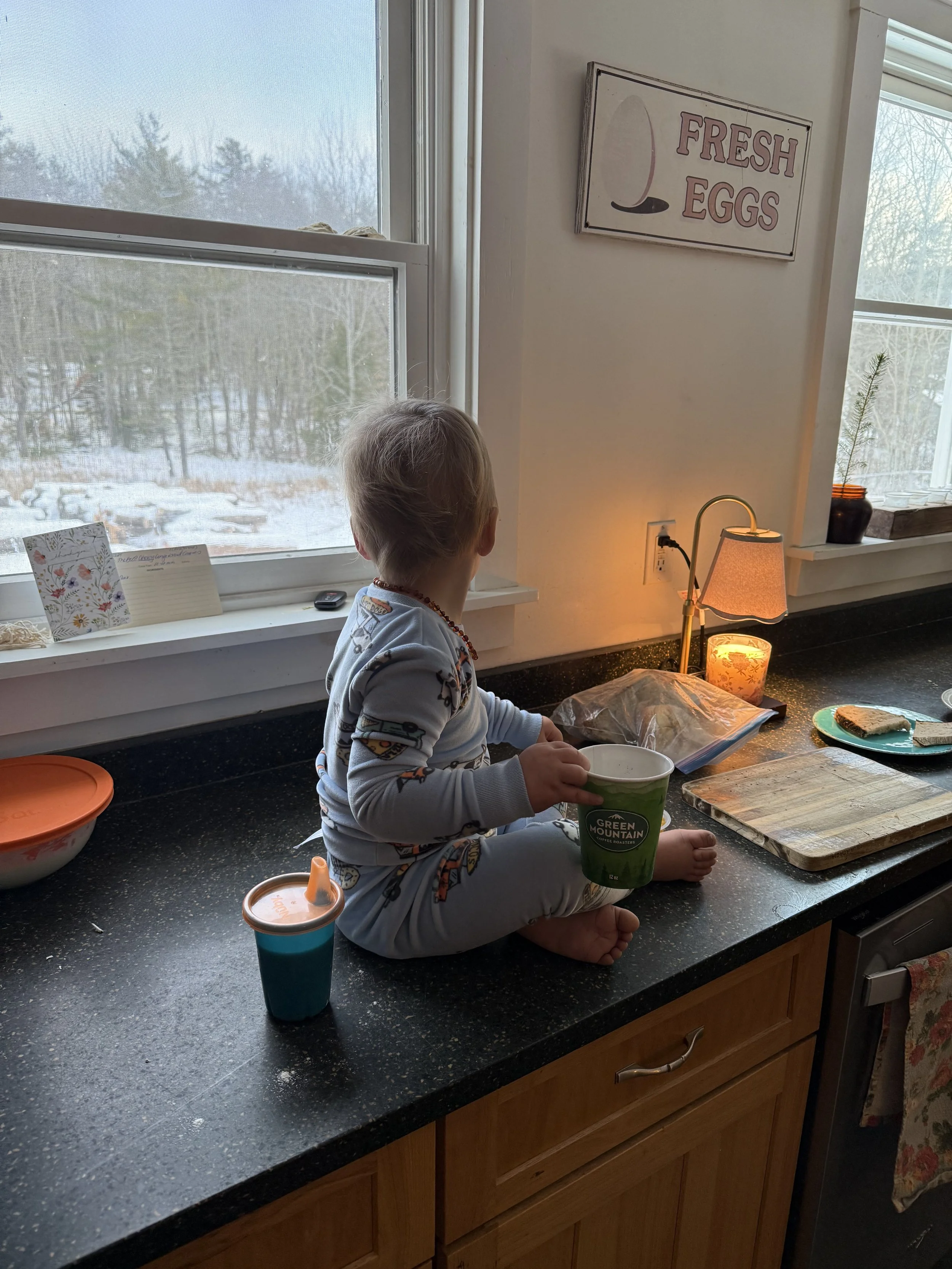 Baby sitting on counter in pjs with a coffee cup by a candle looking outside to snowy winter scene