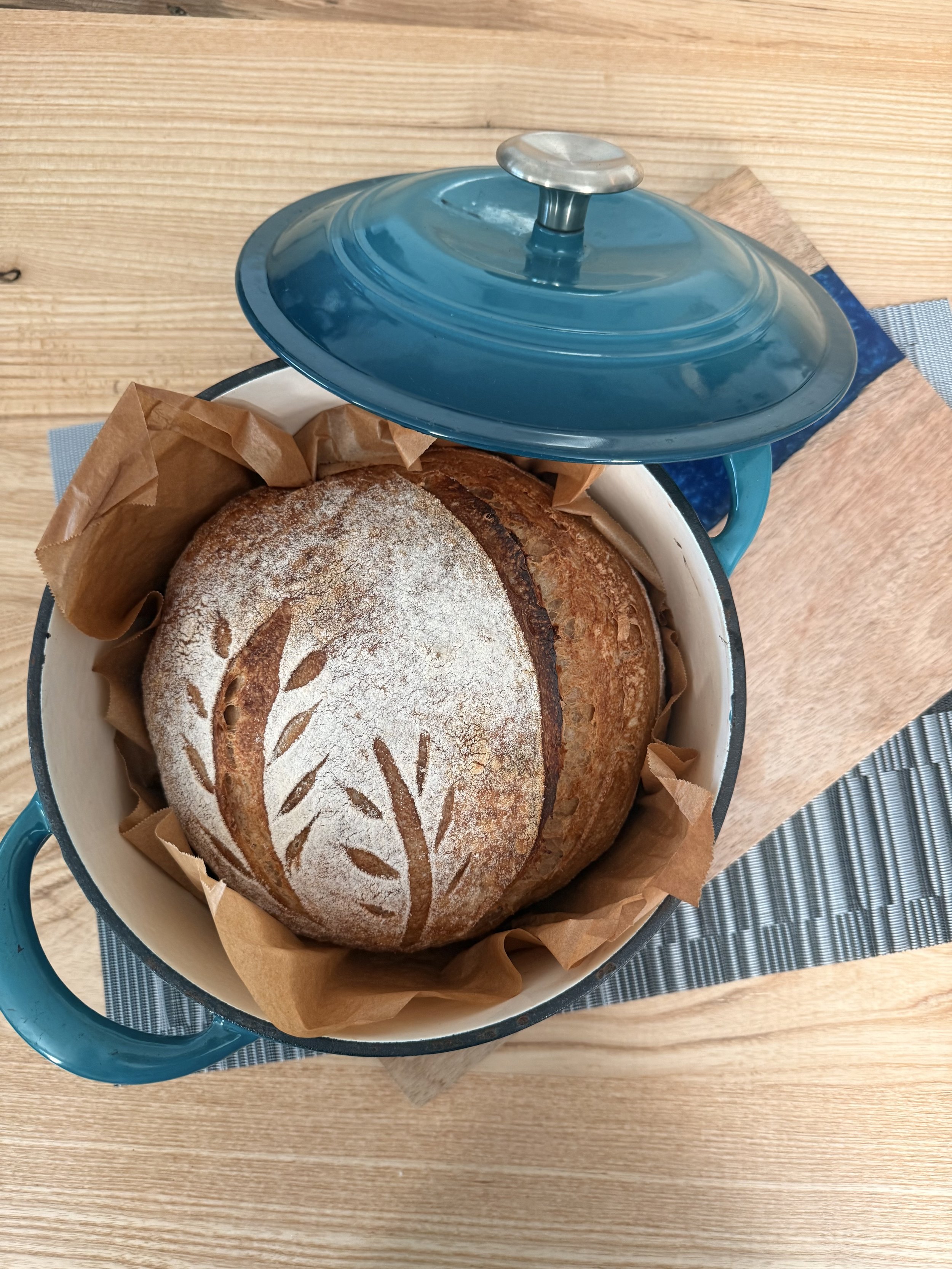 Loaf of bread with a decorative leaf pattern on top, stored in a round container with a blue lid, placed on a wooden surface.