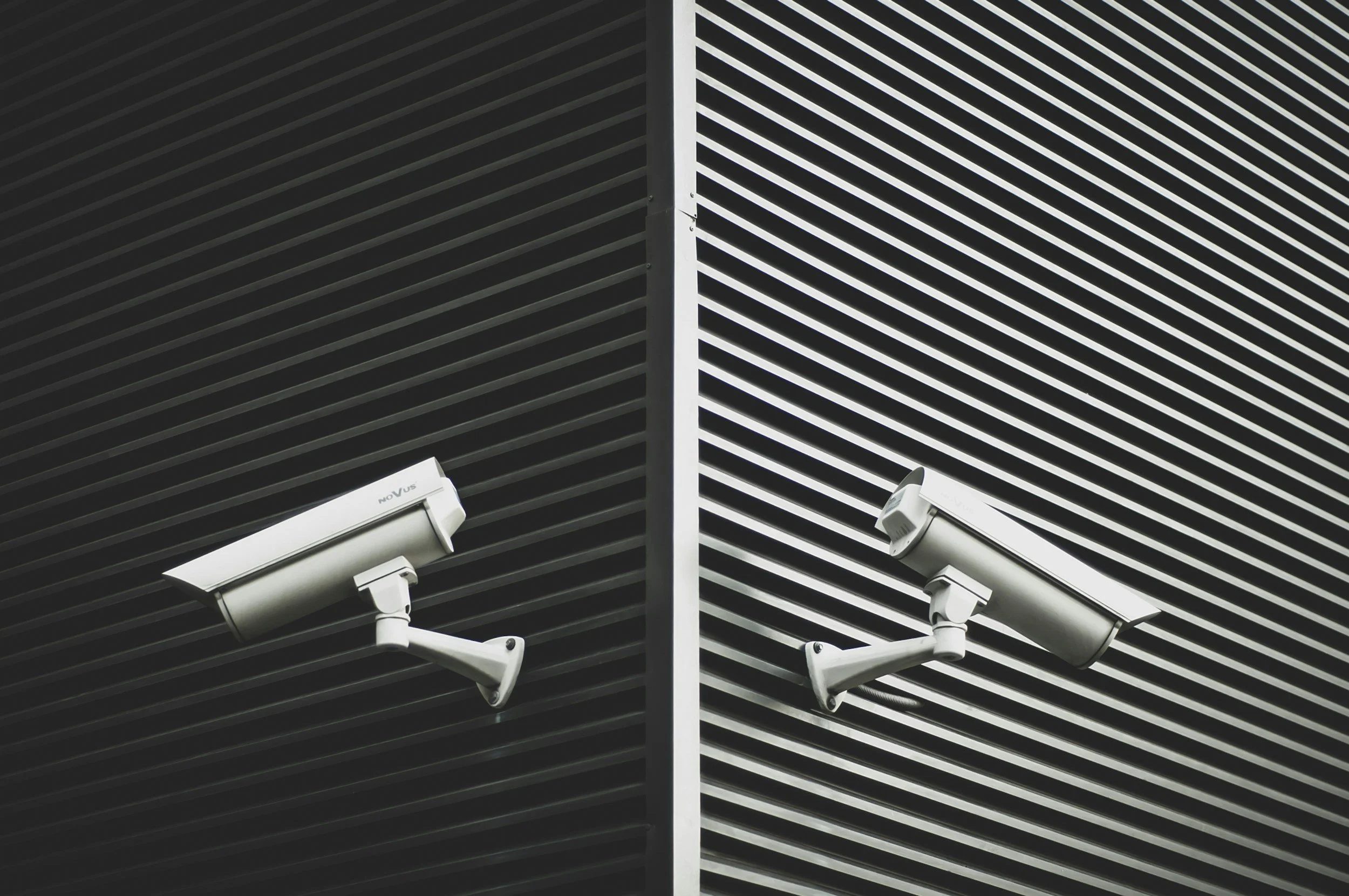 Two security cameras mounted on a building corner, with one on each side, against a background of dark, horizontal metal slats.