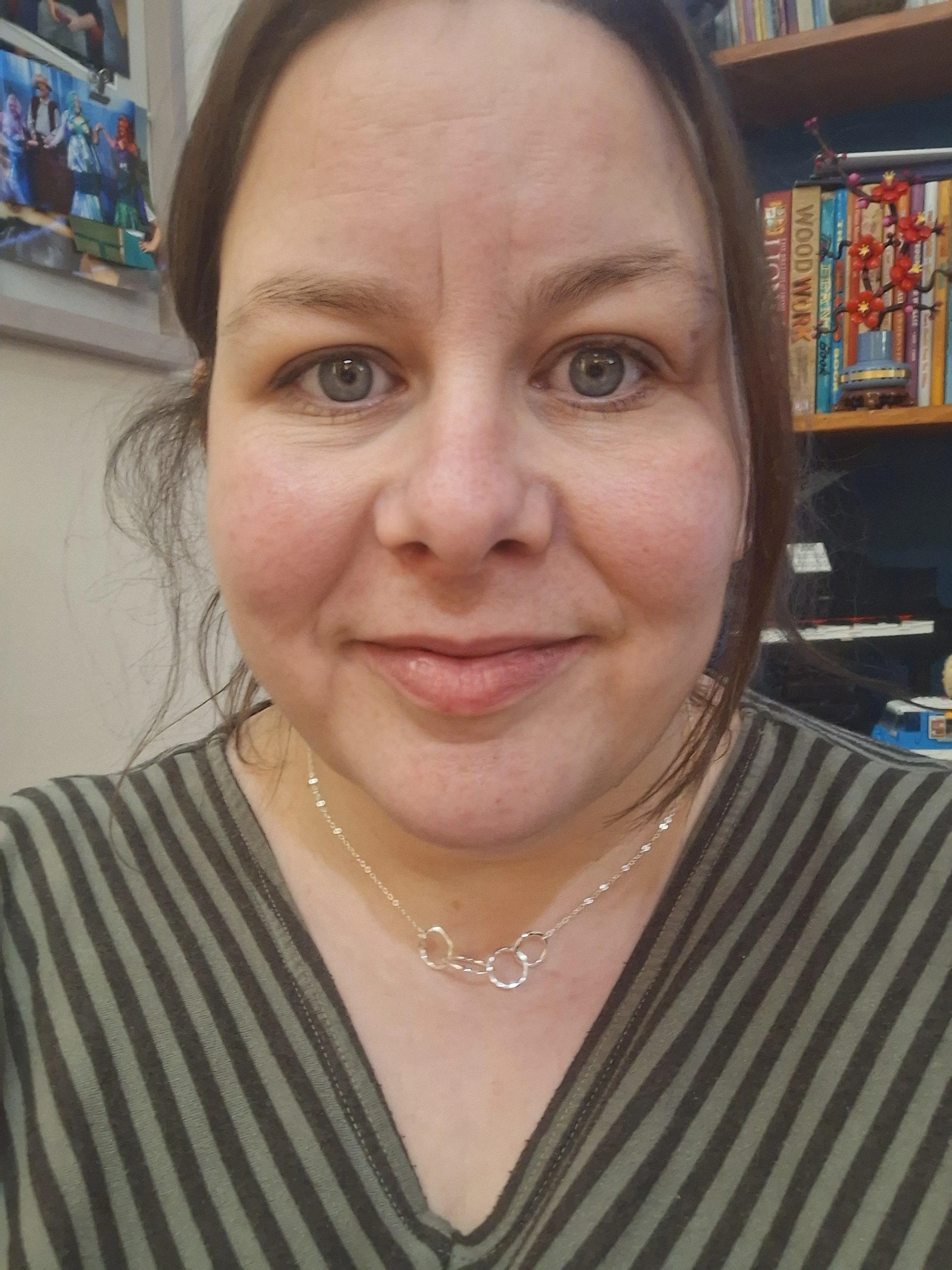 A close-up of a woman smiling, wearing a striped shirt and a silver necklace, with books and toys on shelves in the background.
