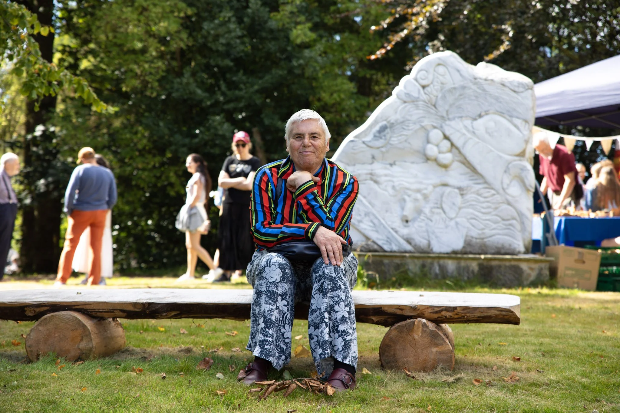 An elderly man with gray hair, wearing a colorful striped shirt and floral pants, sitting on a wooden bench in a park. Behind him, there is a large white sculpture and several people walking and chatting among trees with autumn leaves.