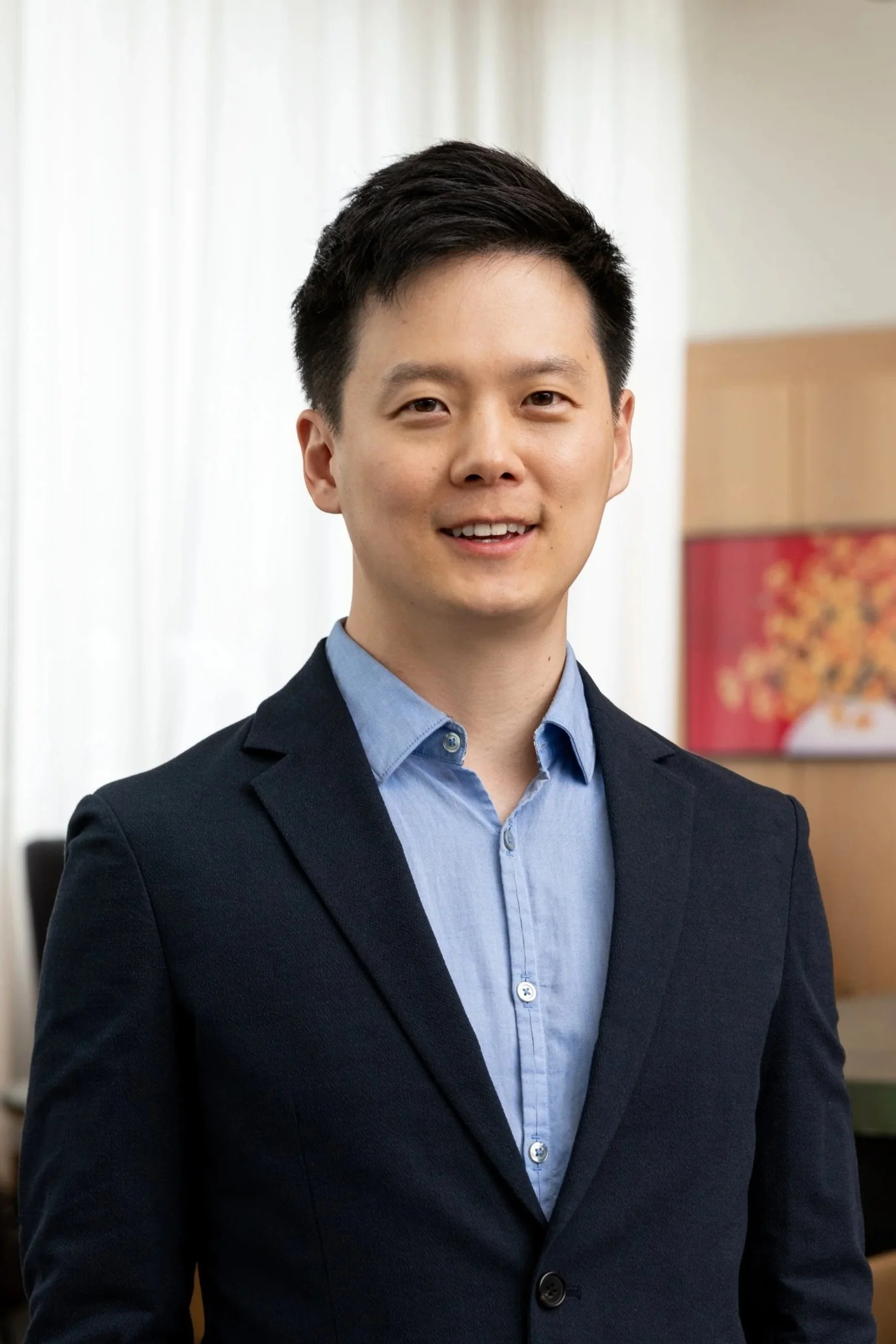 A young man with short black hair, smiling and showing his teeth, wearing a navy blue shirt with small white dots and a grey cable-knit cardigan with a striped trim, against a plain white background.