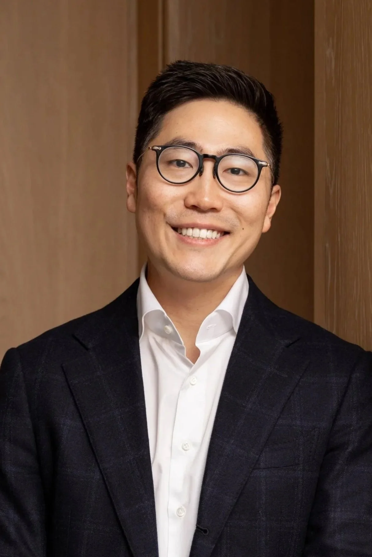 Headshot of a young man with dark hair, glasses, smiling, wearing a white dress shirt and dark blazer against a plain background.