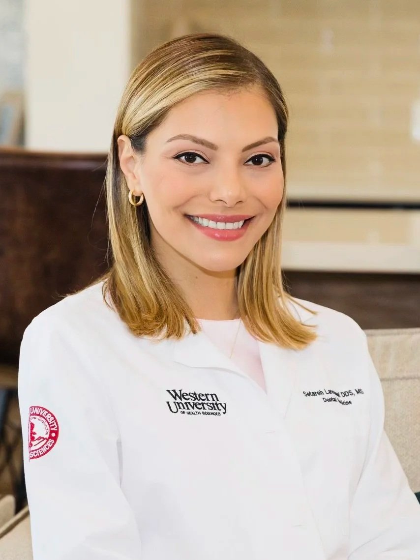 A smiling woman with shoulder-length blonde hair, wearing a white lab coat with 'Western University' and her name embroidered on it, standing indoors.