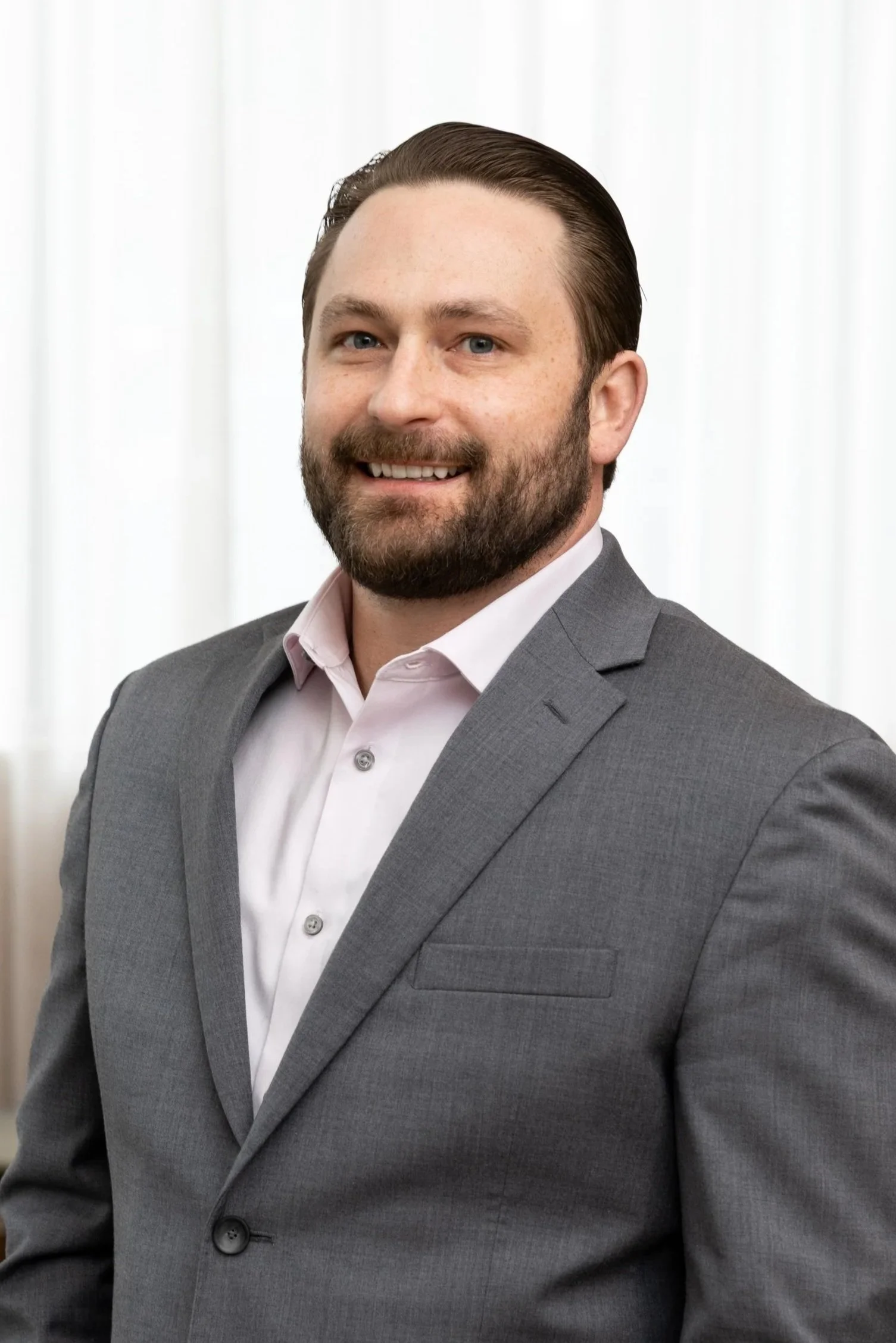 A man with brown hair and a beard, smiling, wearing a teal collared shirt, standing against a light gray background.