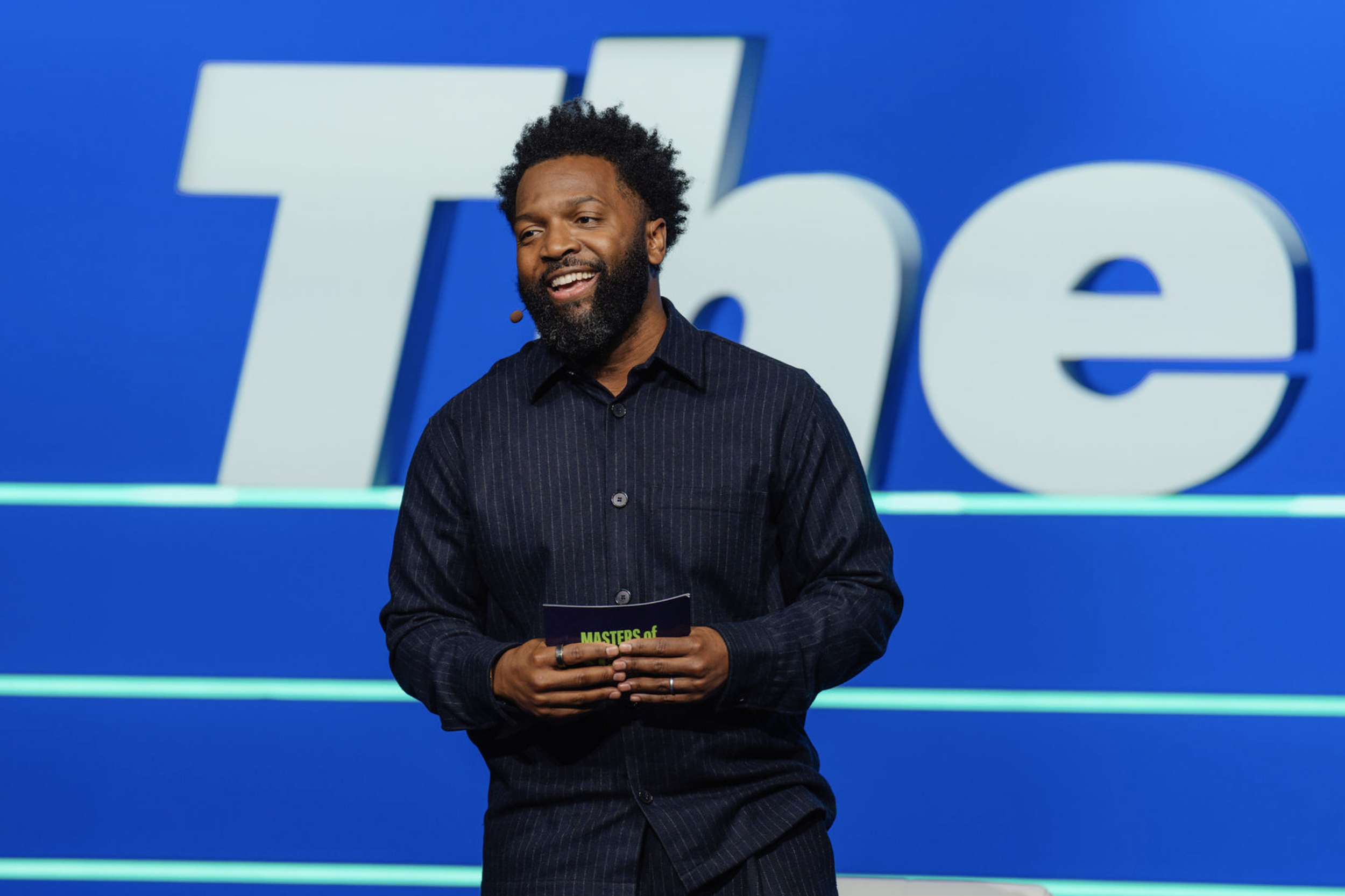 A man standing on stage with a large blue background and the Word 'The' partially visible behind him.