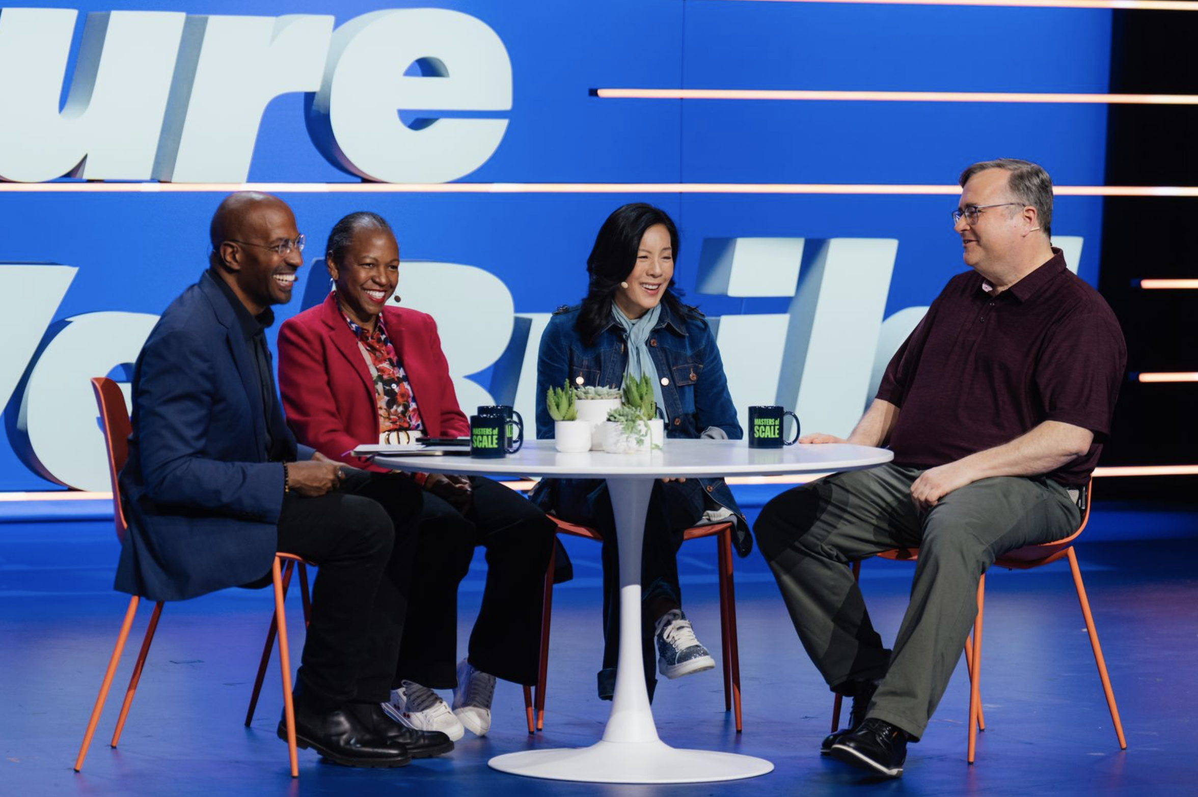 Four people sit around a white round table on a stage, smiling and engaged in conversation. There are two men and two women, with a blue background and large partial text behind them. The table has two black mugs, some plants, and a notebook.