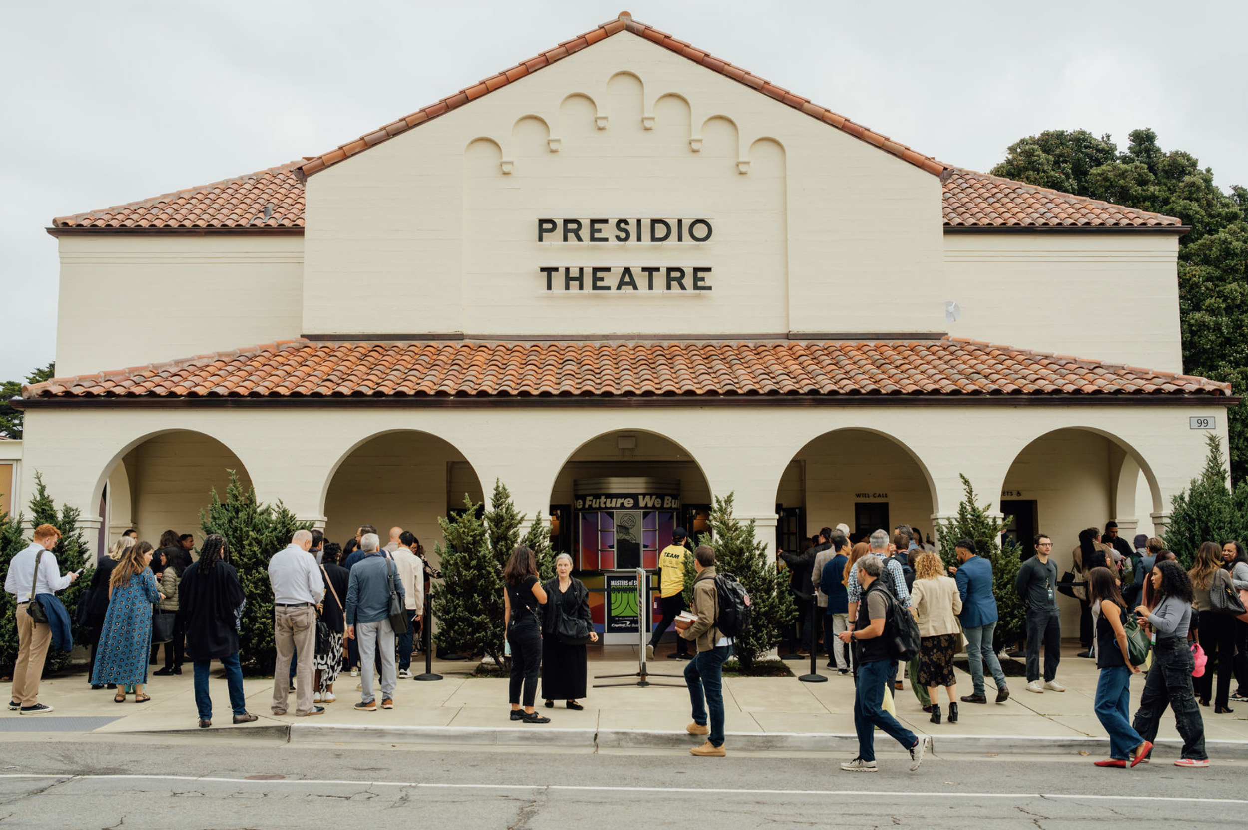 Crowd of people standing outside the Presidio Theatre in San Francisco, California, with trees in the background, and a cloudy sky overhead.