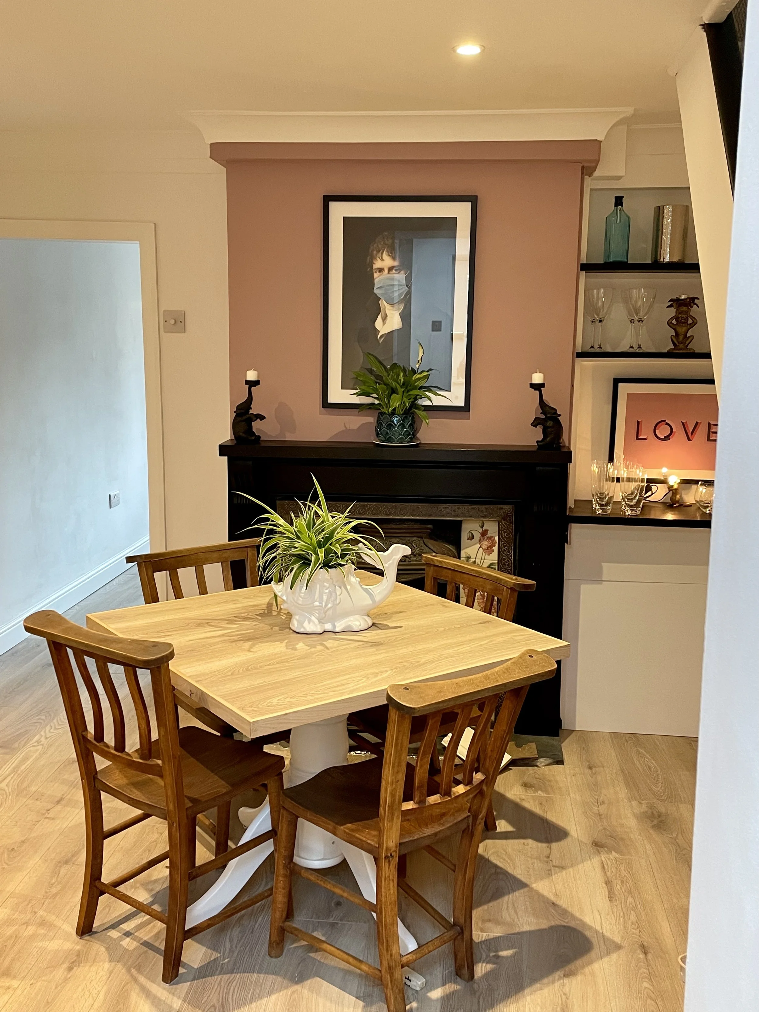 A dining room with a wooden table and four matching chairs, a white ceramic teapot-shaped plant holder on the table, a black fireplace mantel with a framed photo of a person wearing a face mask, a green potted plant, and a decorative shelf with glass