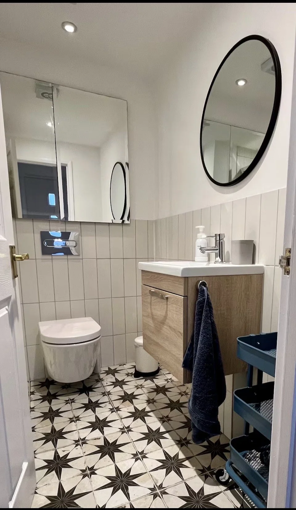 A modern small bathroom featuring a white toilet, a wooden vanity with a sink, an oval mirror, tiled wall, patterned floor tiles, and a small storage cart.
