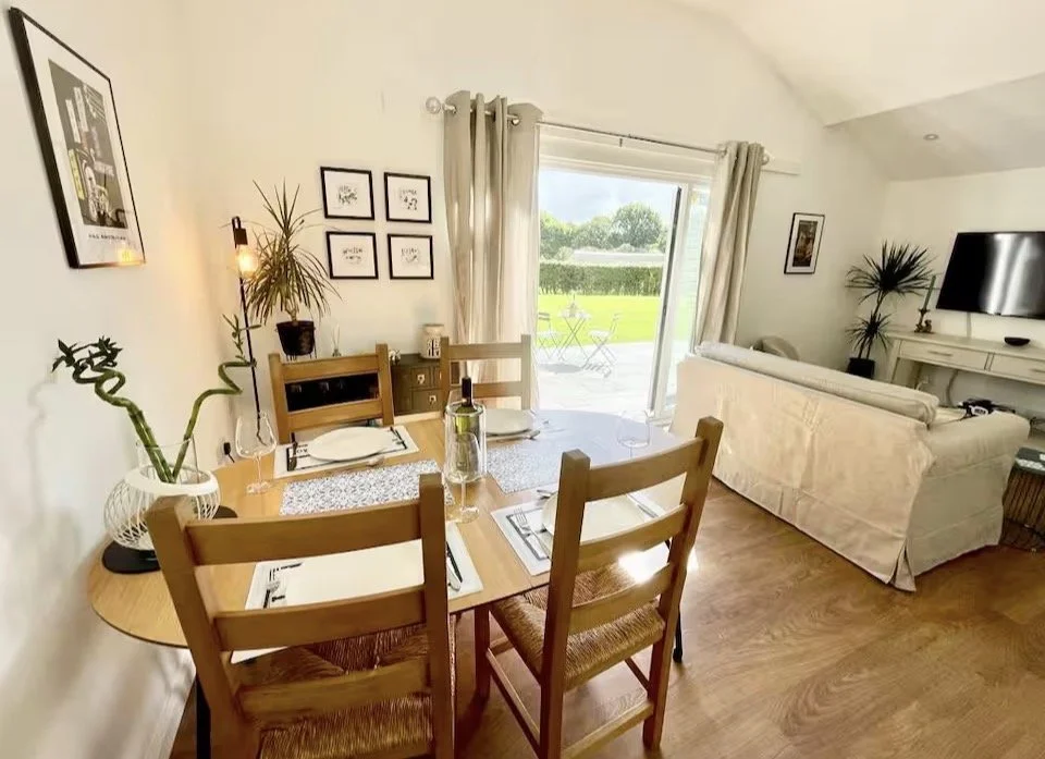 A cozy living and dining area with a wooden dining table set for four, a white couch facing a wall-mounted TV, and a sliding glass door leading to a green outdoor lawn with trees and a small patio.