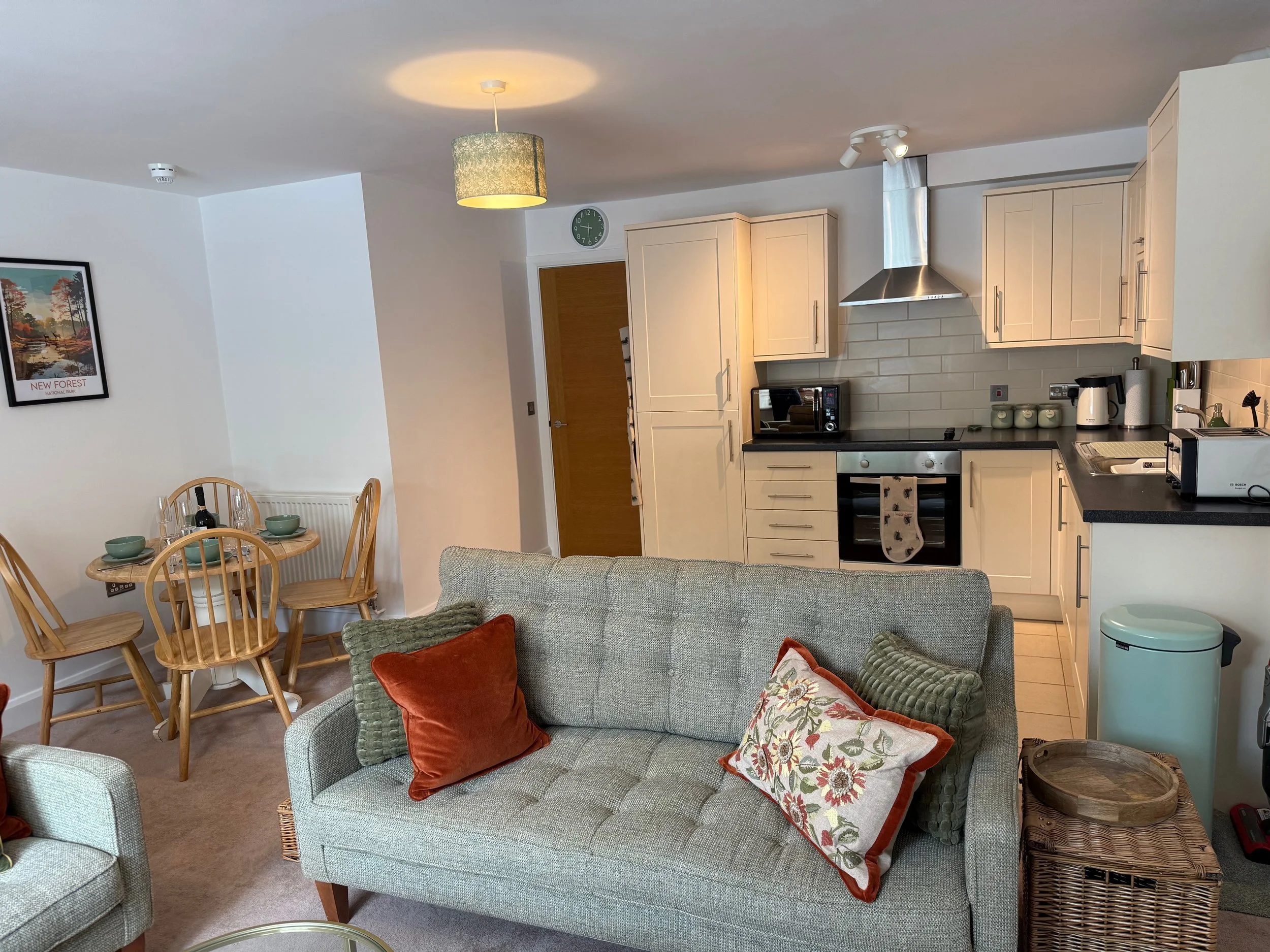 Living room with a beige sofa decorated with orange and green pillows, adjacent to an open kitchen with light cabinets, black countertops, and stainless steel appliances. There is a small dining table with four wooden chairs nearby.