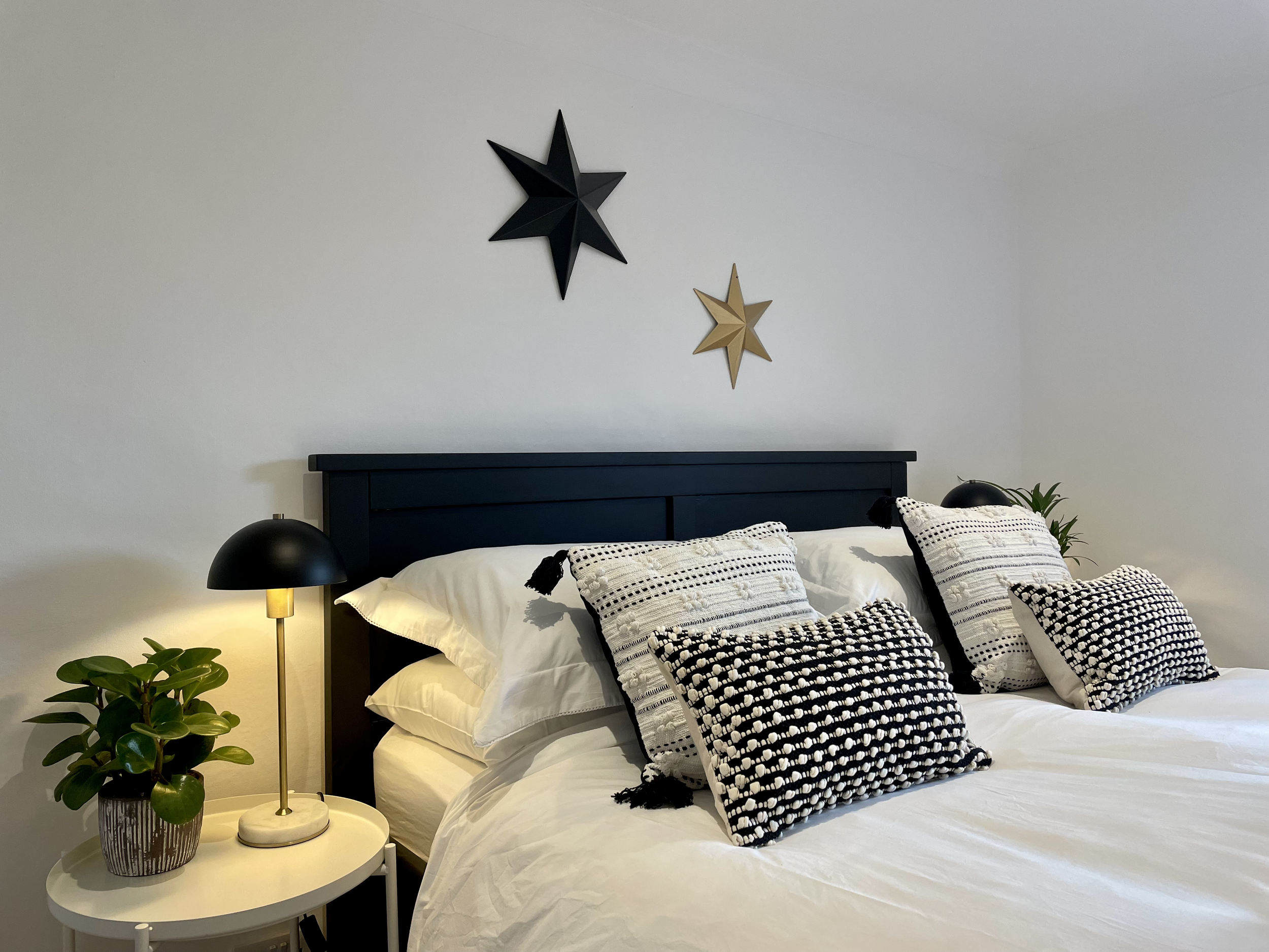 A neatly made bed with white bedding and various black and white patterned pillows, a navy blue headboard, two black bedside lamps, a white bedside table with a small potted plant, and two decorative star wall hangings in black and gold on a plain wh