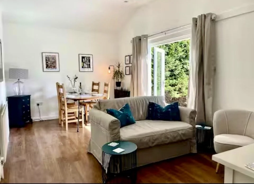 Living room with beige sofa, white armchair, wooden dining table with chairs, large window with open curtains showing greenery outside, side tables, framed pictures on white walls, wooden floor.