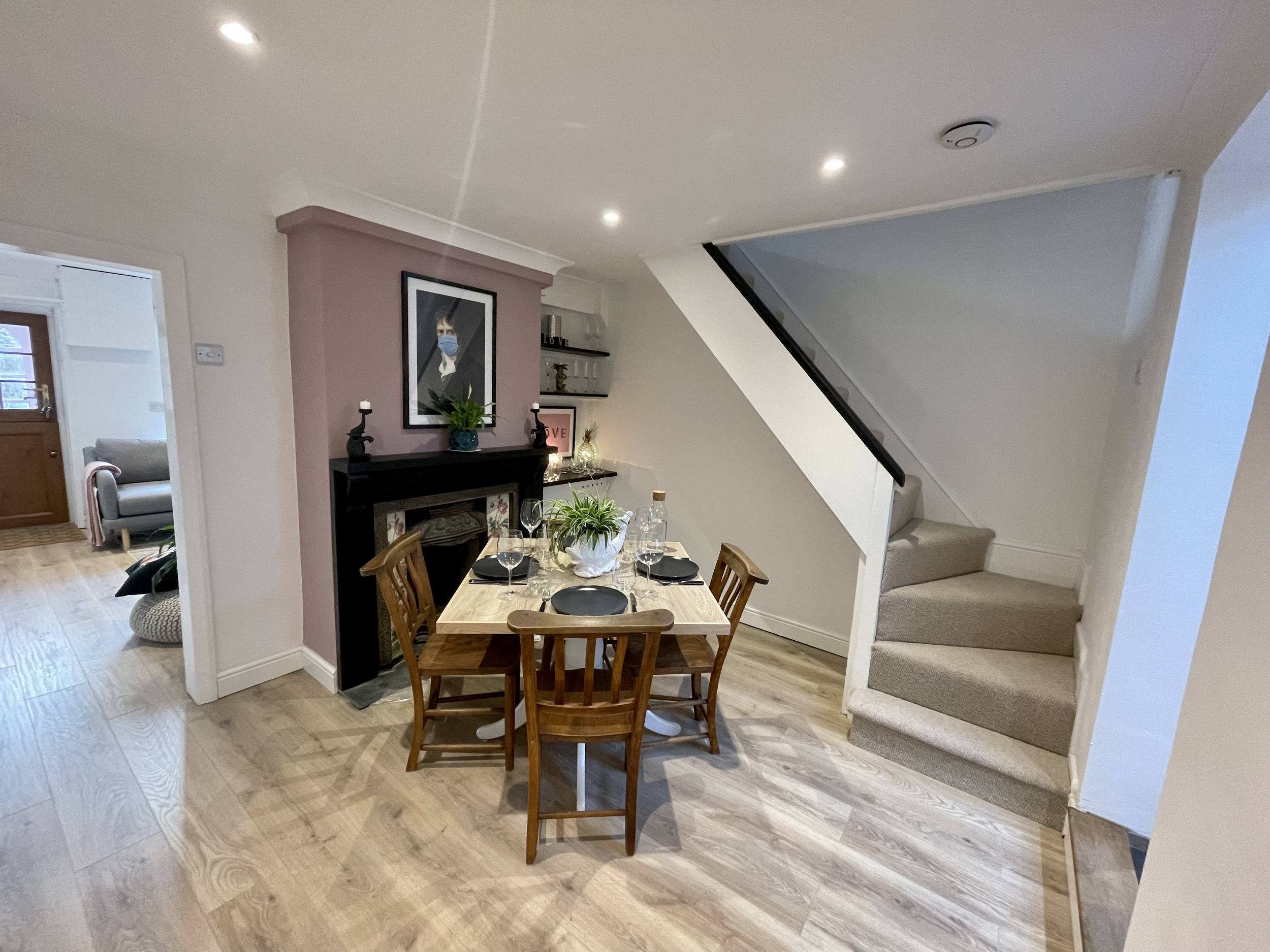 Dining area with a wooden table set with black plates, glasses, and a white vase with green plants. A staircase with beige carpet leads upstairs to the right, and the wall behind the dining table has a purple accent with framed artwork and decorative