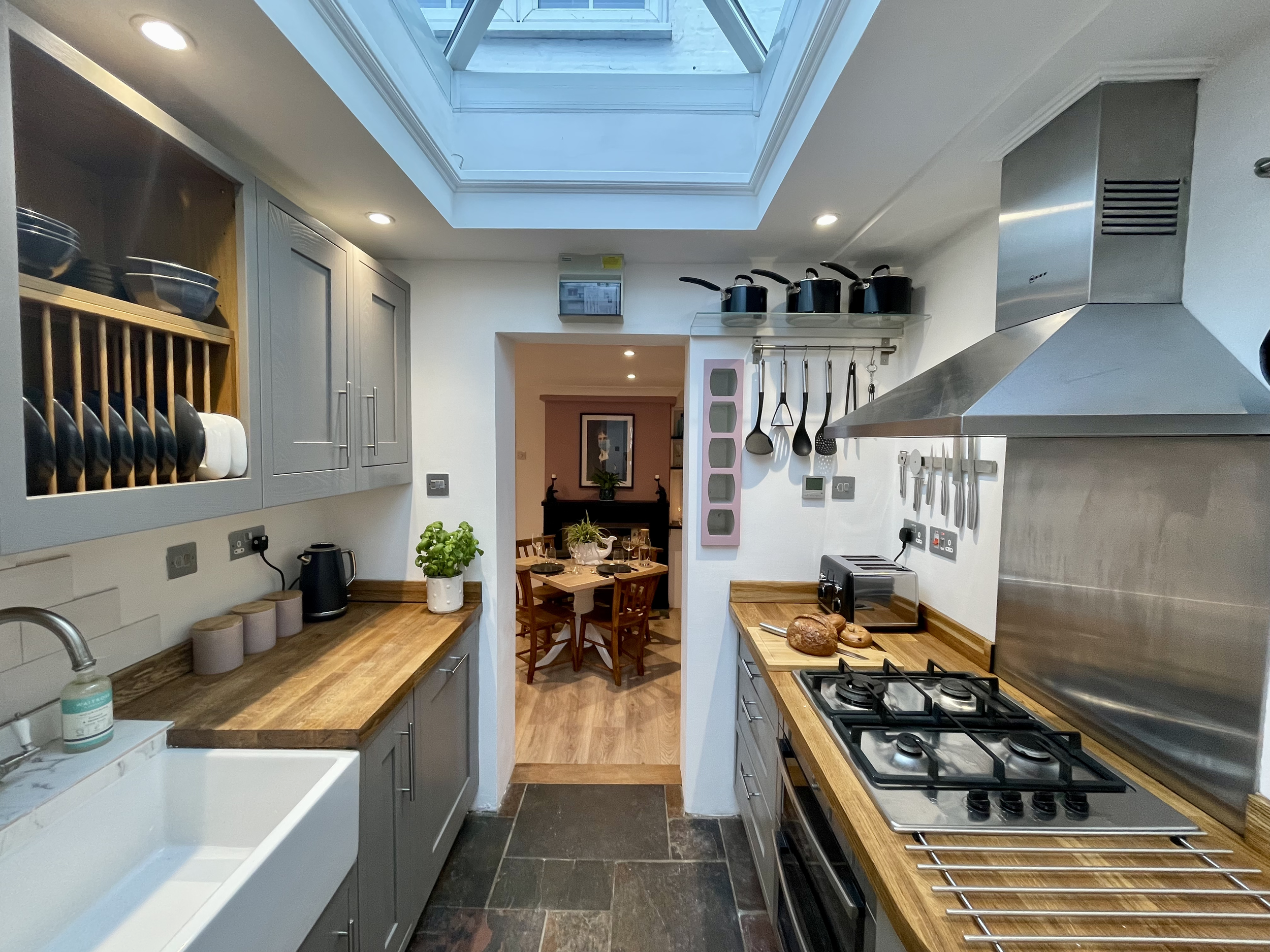 A modern kitchen with gray cabinets, a wooden countertop, and a skylight on the ceiling. There are pots on a shelf above and various utensils hanging on the wall. The stove with a stainless steel backsplash is on the right, and a dining area is visib