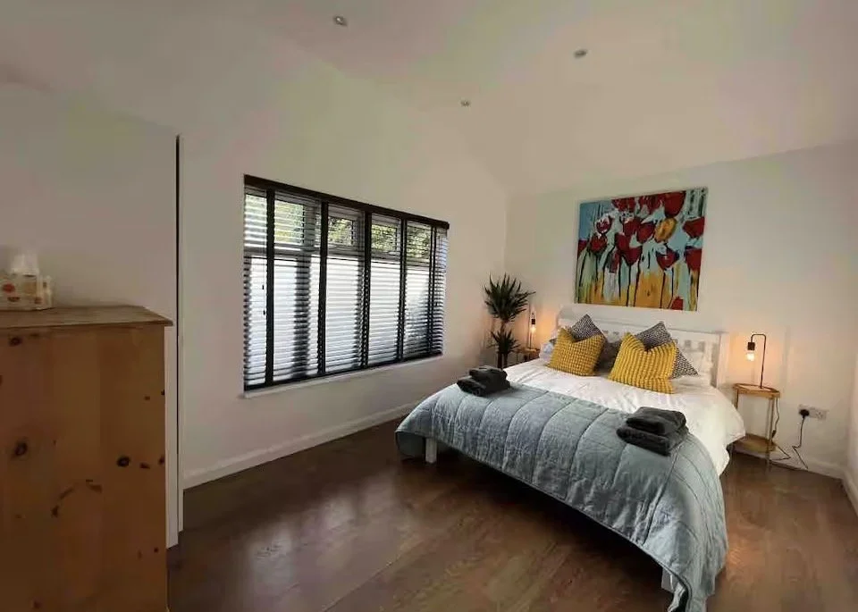 A bedroom featuring a bed with a grey quilt, yellow and black pillows, and artwork of flowers above the headboard. There are two small bedside tables with lamps on each side of the bed, a tall plant near the window, and a wooden dresser on the left. 