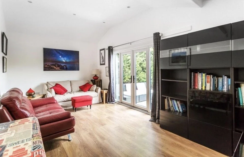 Living room with beige and red sofas, a black bookshelf, and a large window with sliding doors leading to an outdoor space.