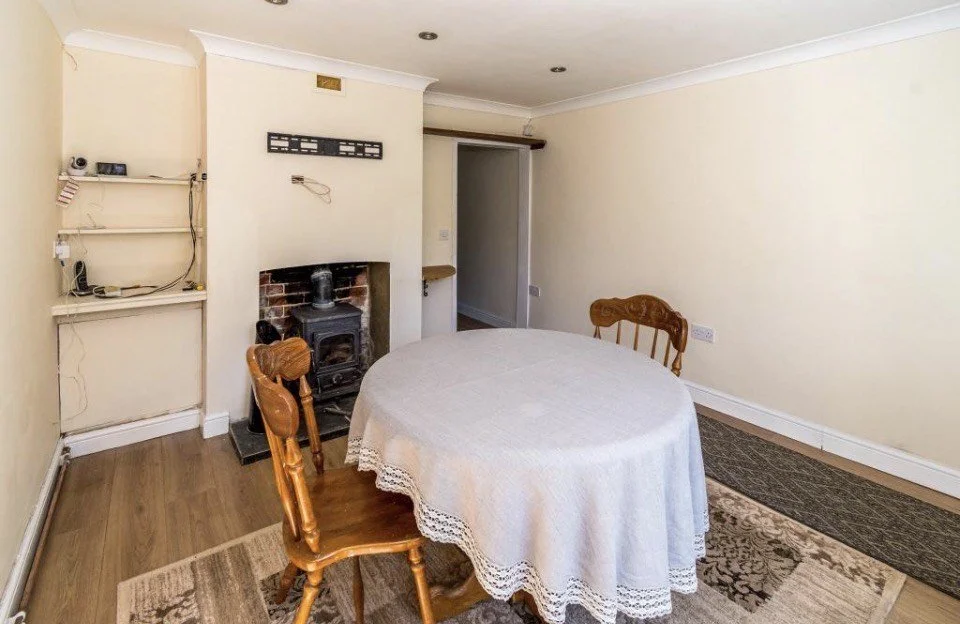 Living room with a round table covered with a white tablecloth and three wooden chairs, a brick fireplace with a wood stove, empty shelves, and a beige wall.