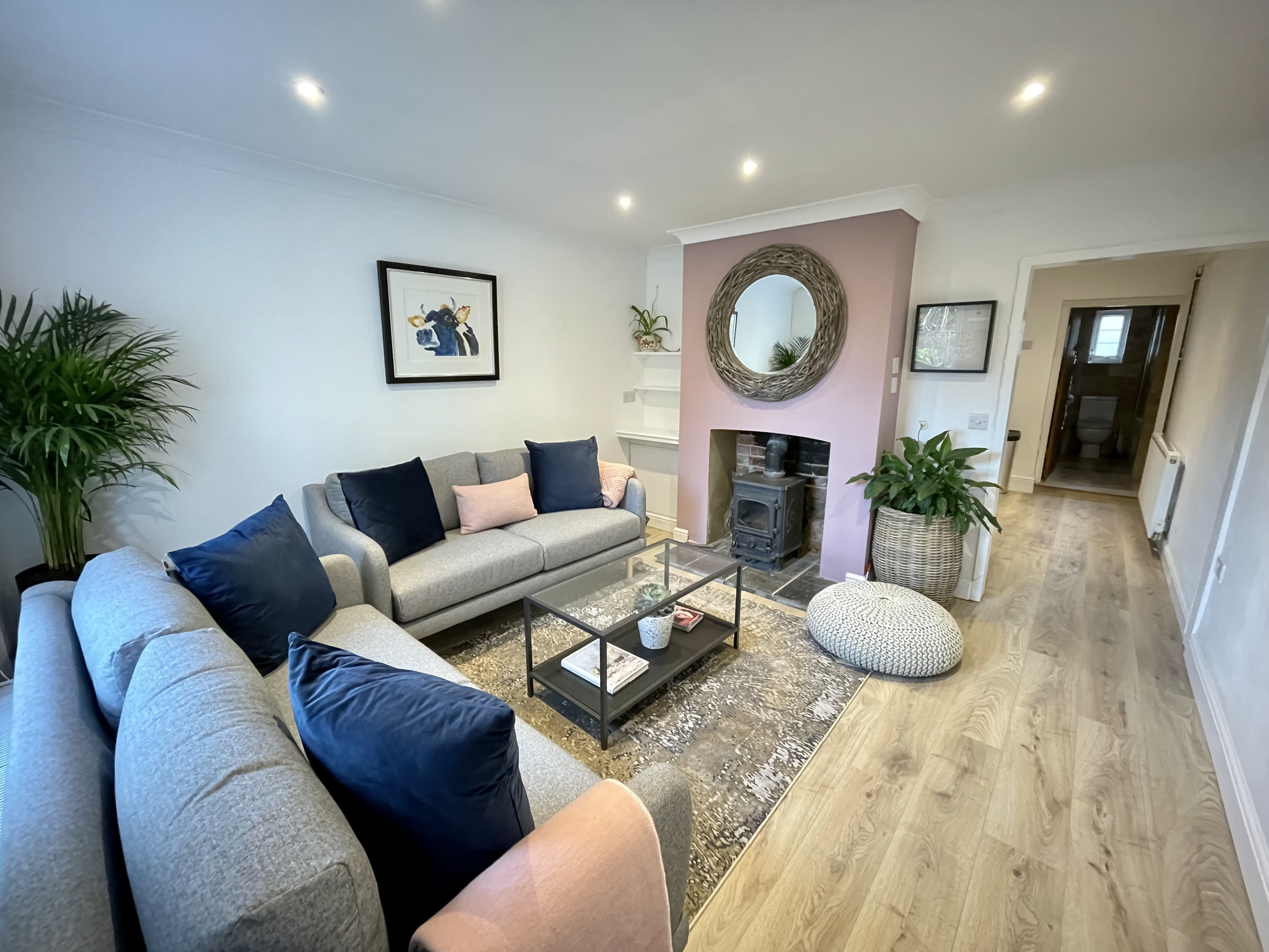 Living room with two beige sofas, navy and light pink cushions, a glass coffee table, a patterned pouf, a potted plant, a decorative mirror above a pink feature wall, a wood stove, and a framed artwork of a cow.