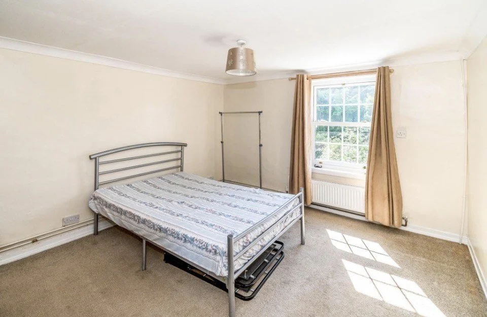Empty bedroom with a metal bed frame, a mattress, a window with beige curtains, and a ceiling light fixture.