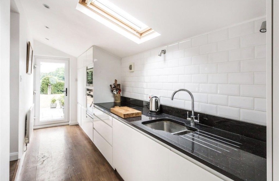 Modern kitchen with white cabinets, black granite countertop, stainless steel sink and faucet, white tile backsplash, a wooden cutting board, and a toaster. Skylight above and a door leading outside.