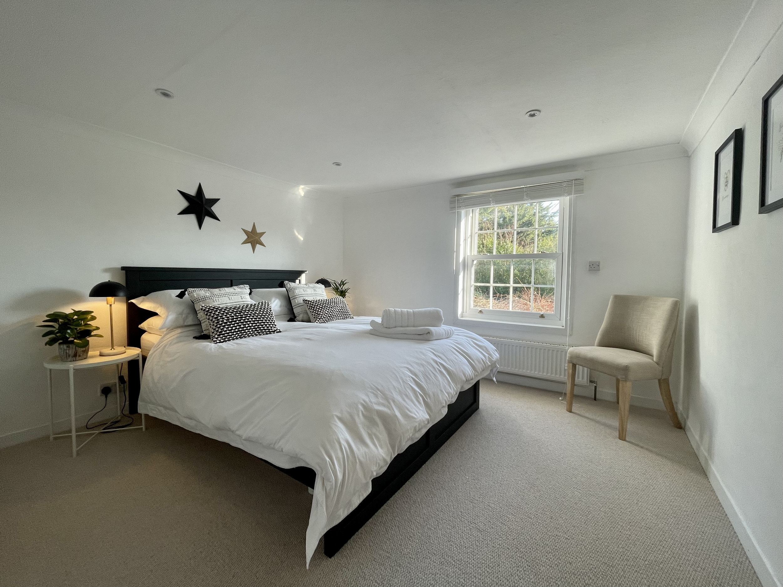 A neatly made bed with white bedding and black and white pillows, with a towel and rolled blanket on top, in a bright bedroom with a window showing greenery outside, beige carpet, a white side table with a potted plant and lamp, and a beige chair nea