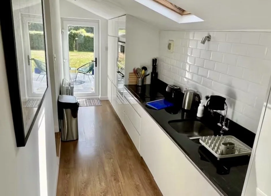 Kitchen with white cabinets, black countertop, and white subway tile backsplash, featuring a sink, dish drying rack, and small appliances, with a sliding glass door leading to an outdoor patio with chairs and a lawn.