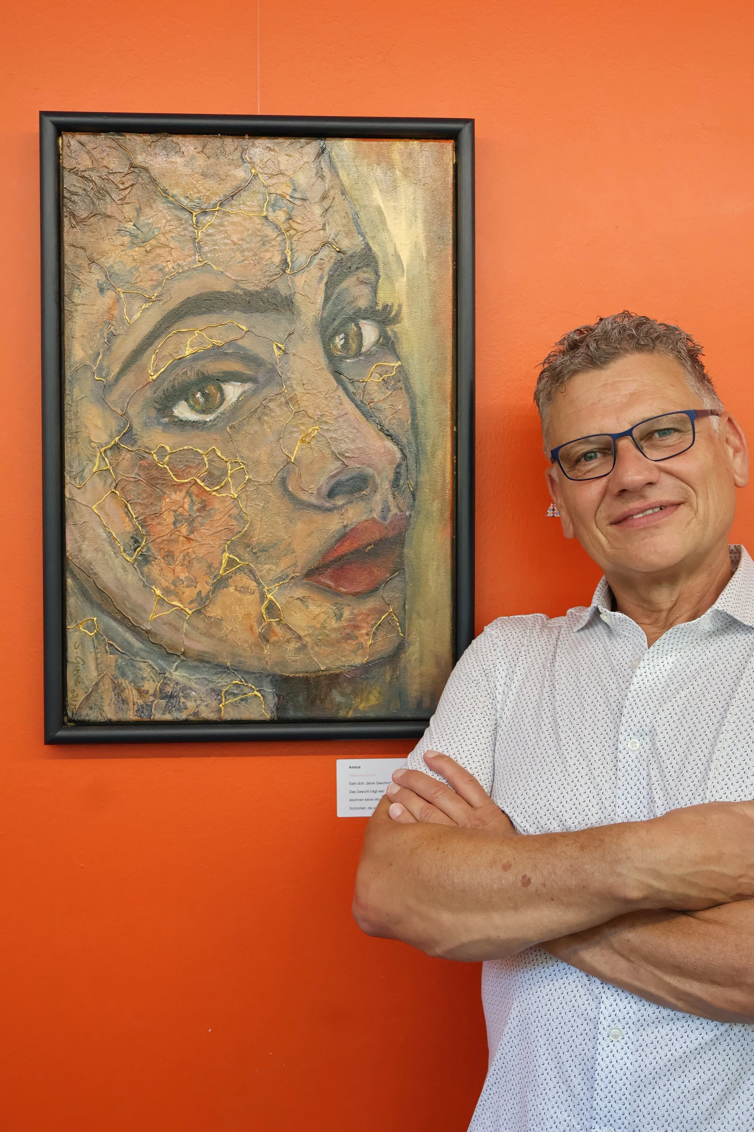 Ein Mann mit Brille und Kurzhaar trägt ein weißes Hemd und steht vor einem orangefarbenen Wand hinter einem Gemälde einer weiblichen Figur mit heiliger Erscheinung, das im Kunststil realistisch gehalten ist und Goldakzente aufweist.